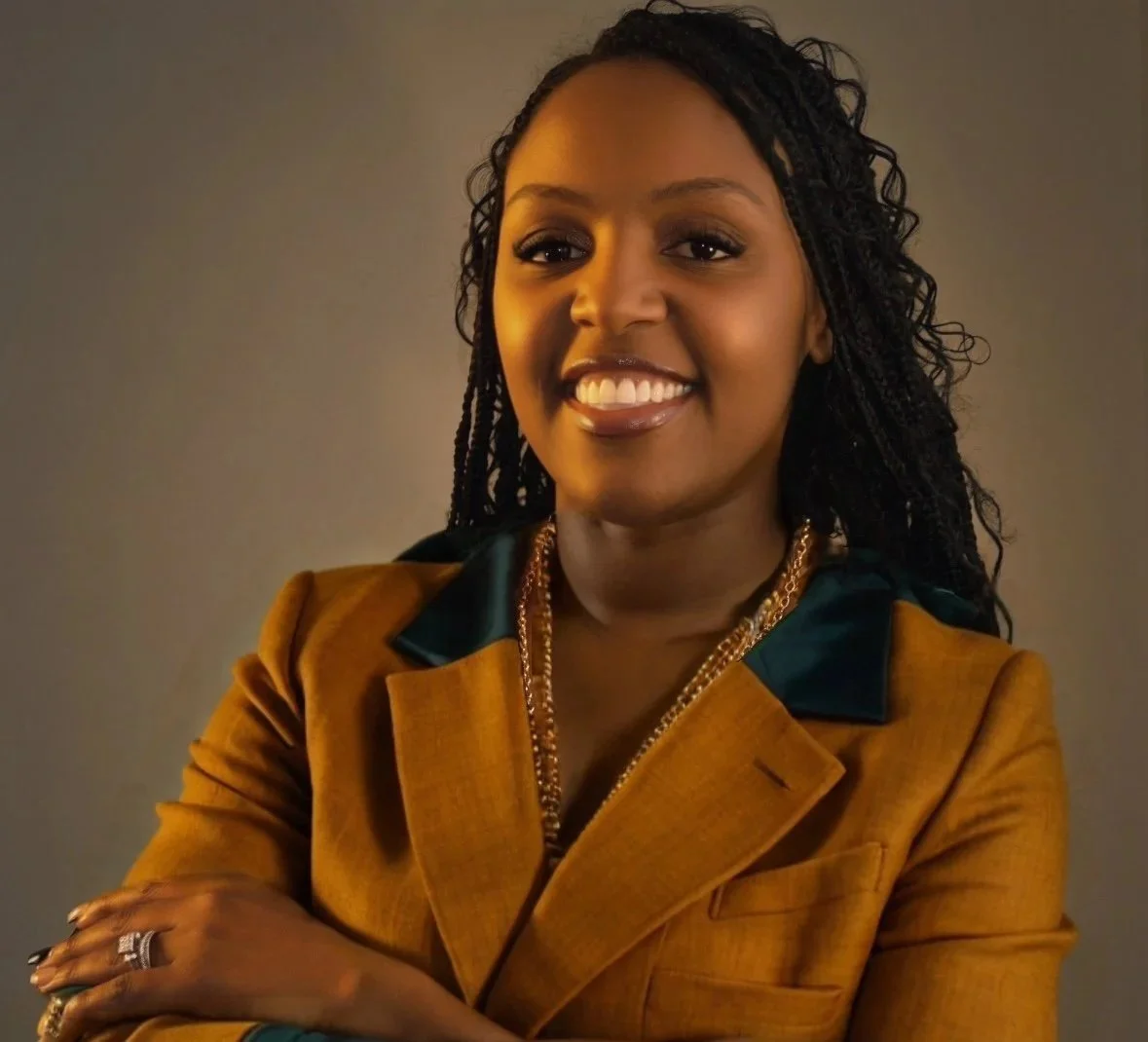 A woman with dark curly hair, smiling, wearing a mustard-colored blazer with a dark collar, accessorized with gold necklaces and rings, posing with crossed arms against a plain background.