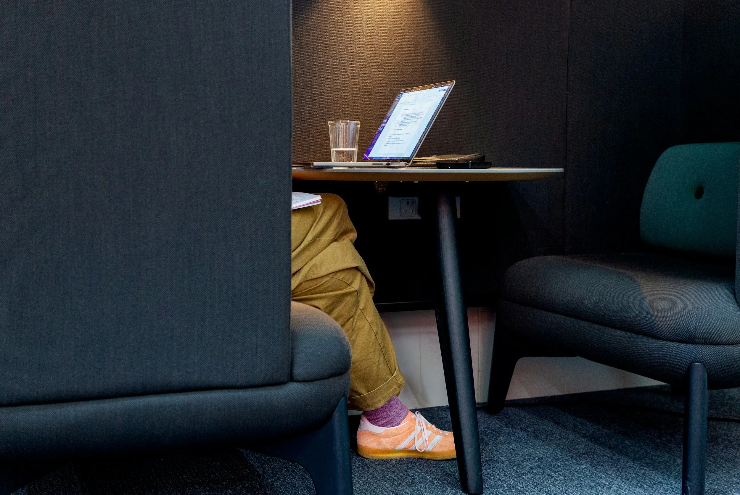 Part of a person's legs wearing yellow pants and orange shoes are visible under a table with a glass of water and a laptop. The person is seated between two dark-colored cushioned chairs in a small office or study space.