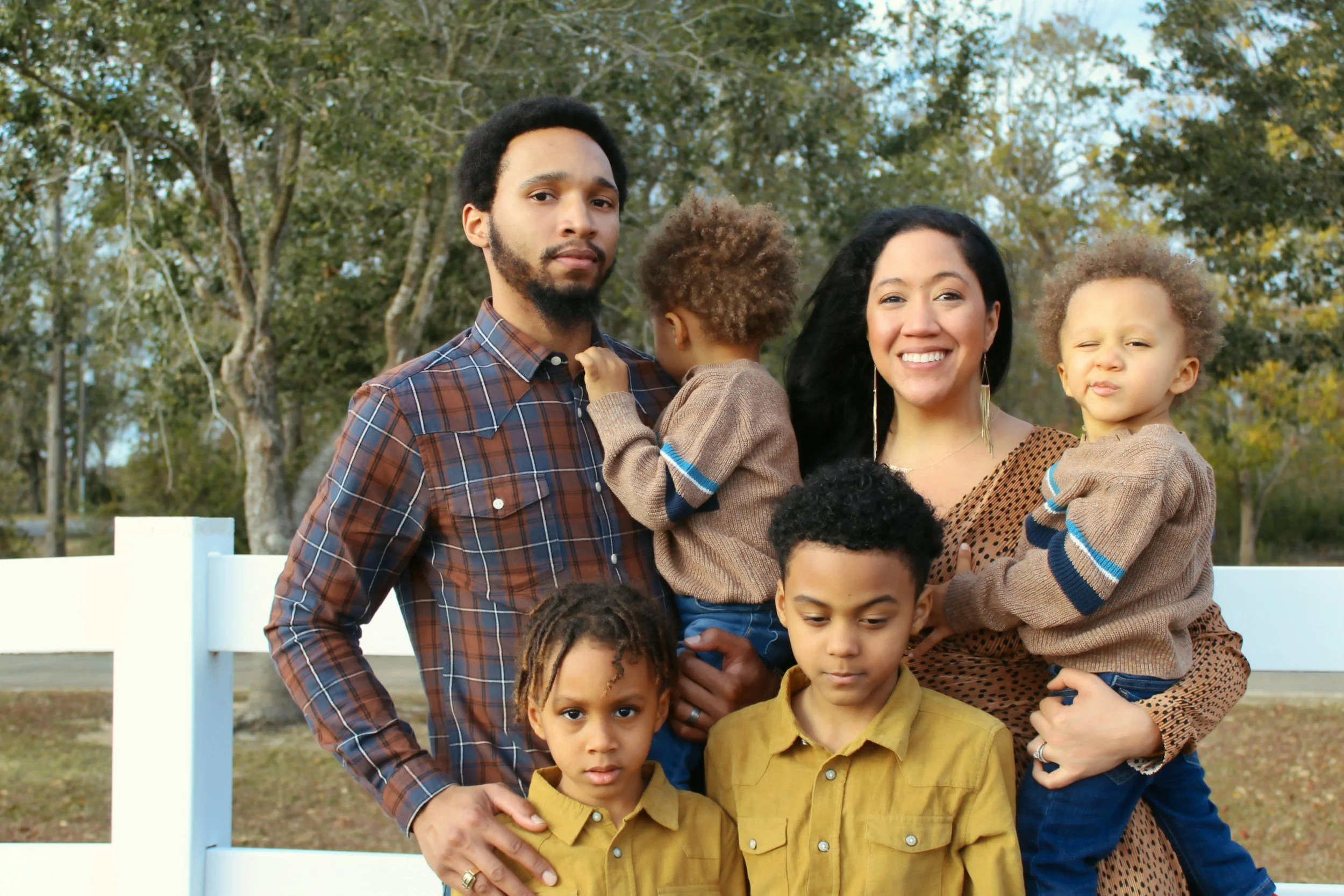 A family of six standing outdoors in front of a white fence and trees. The father, with a beard, holds one child, and the mother, smiling, holds another child. Two young boys stand in front, one with a serious expression and the other with a smirk. All are dressed casually in warm clothing.