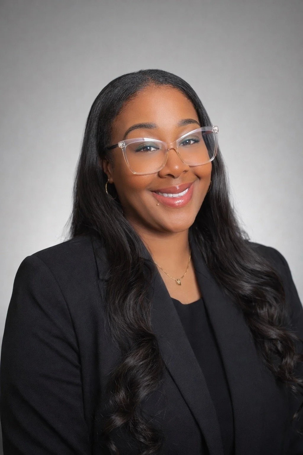 Portrait of a smiling woman with long dark hair, wearing glasses, a black blazer, and jewelry, against a plain light-colored background.