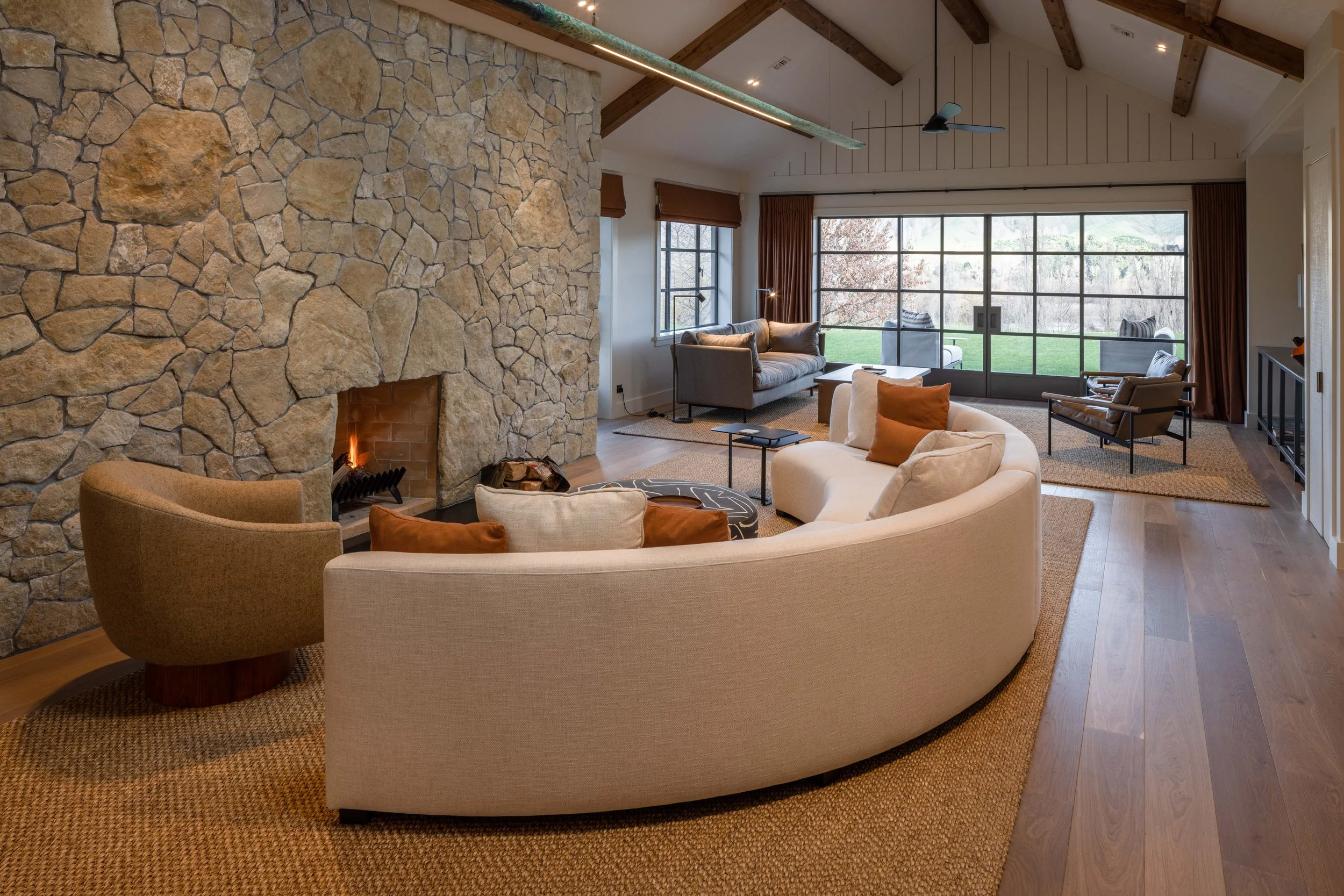 Living room with a stone fireplace, curved beige sofa, armchair, and various other seating options, large windows showing a grassy outdoor area.