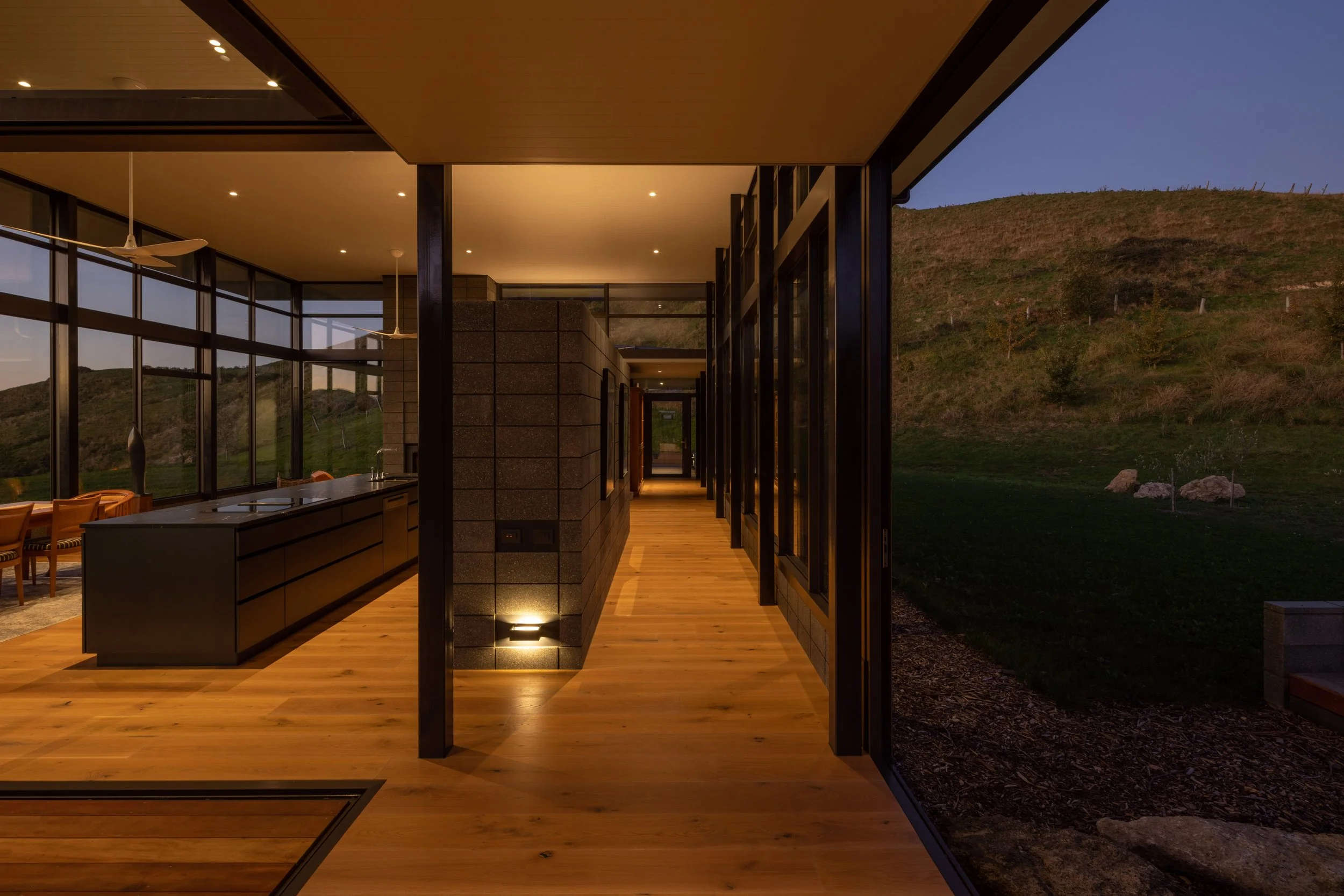 Interior of a modern house with wooden floors, black accents, and large glass windows overlooking a hillside at dusk.