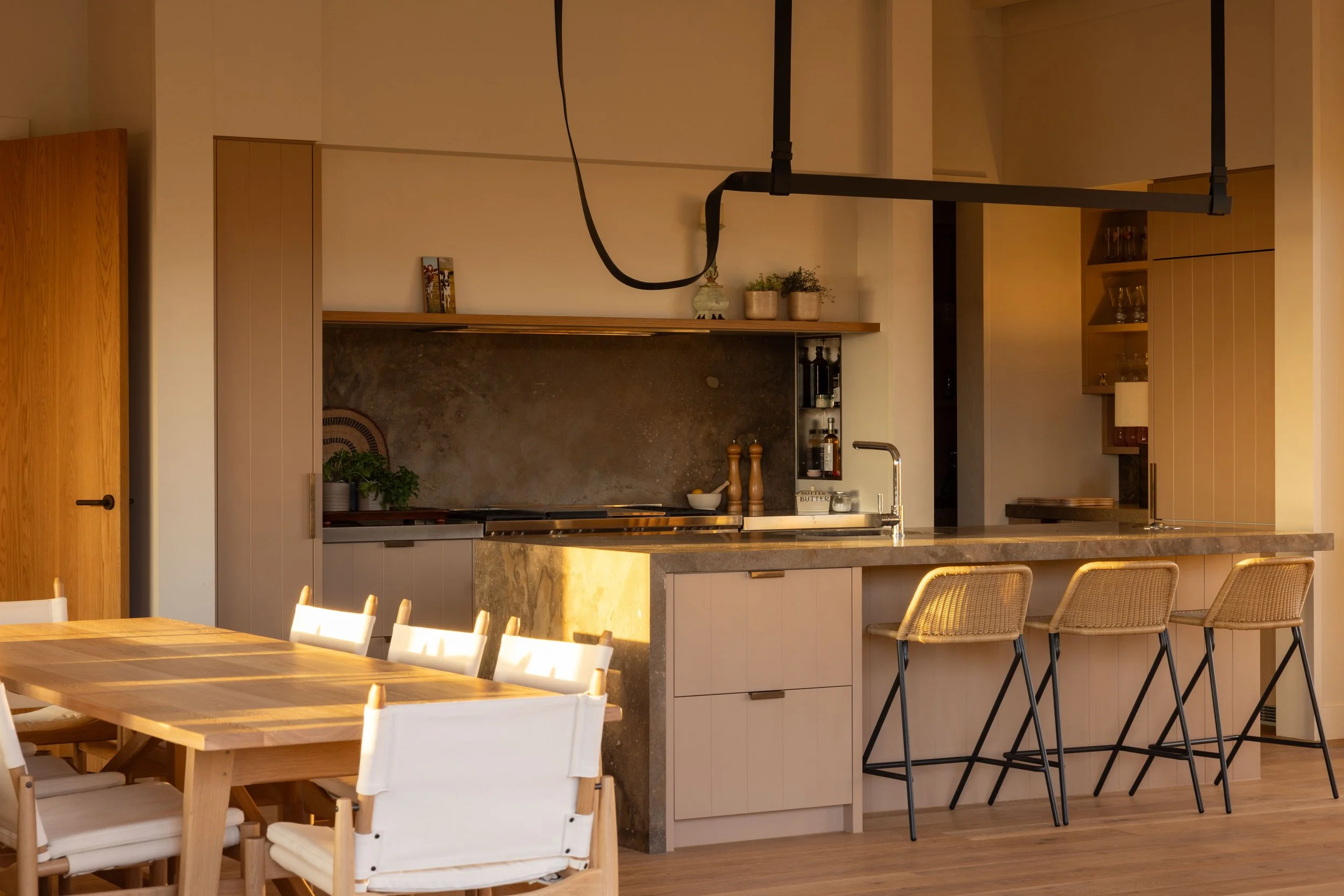 Modern kitchen with a marble island, beige cabinets, a black metal pot rack hanging from the ceiling, and a dining table with white chairs. Sunlight casts a warm glow on the space.