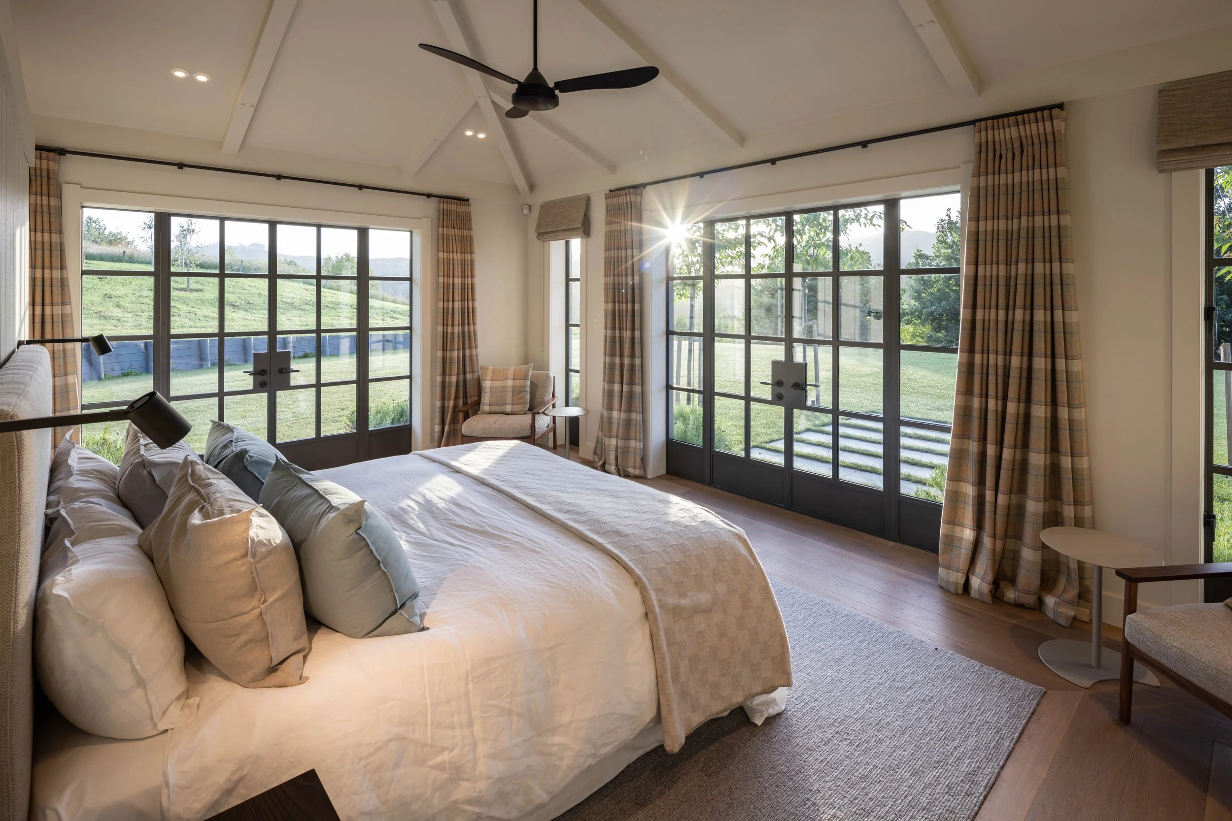 Bright bedroom with large windows and sliding glass doors overlooking a green landscape, ceiling fan, cozy bed with multiple pillows, and seating area.