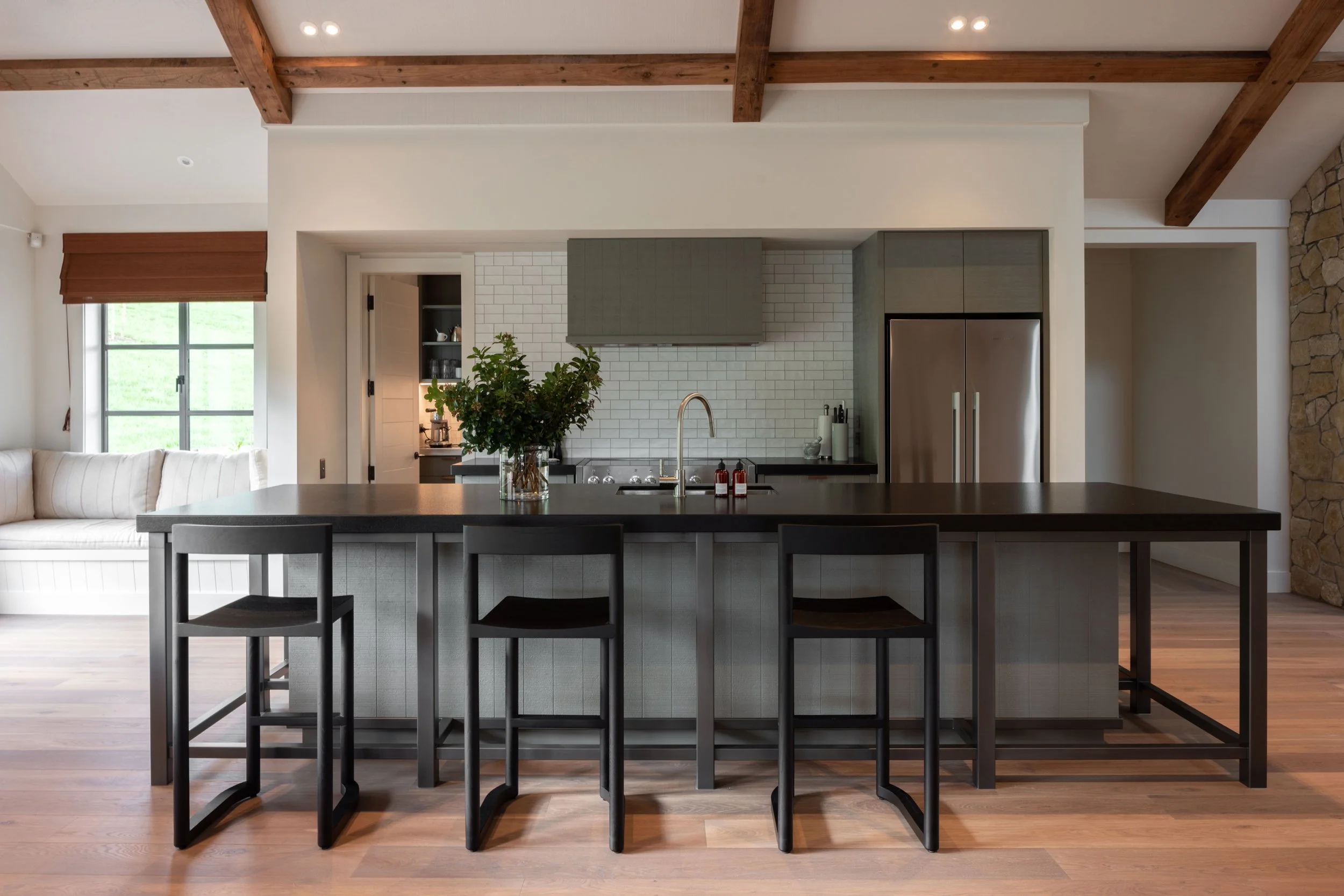 Modern kitchen with a large black island counter, three black chairs, a stainless steel refrigerator, white tile backsplash, gray cabinets, and a window with a wooden blind. There's a vase with greenery on the island.