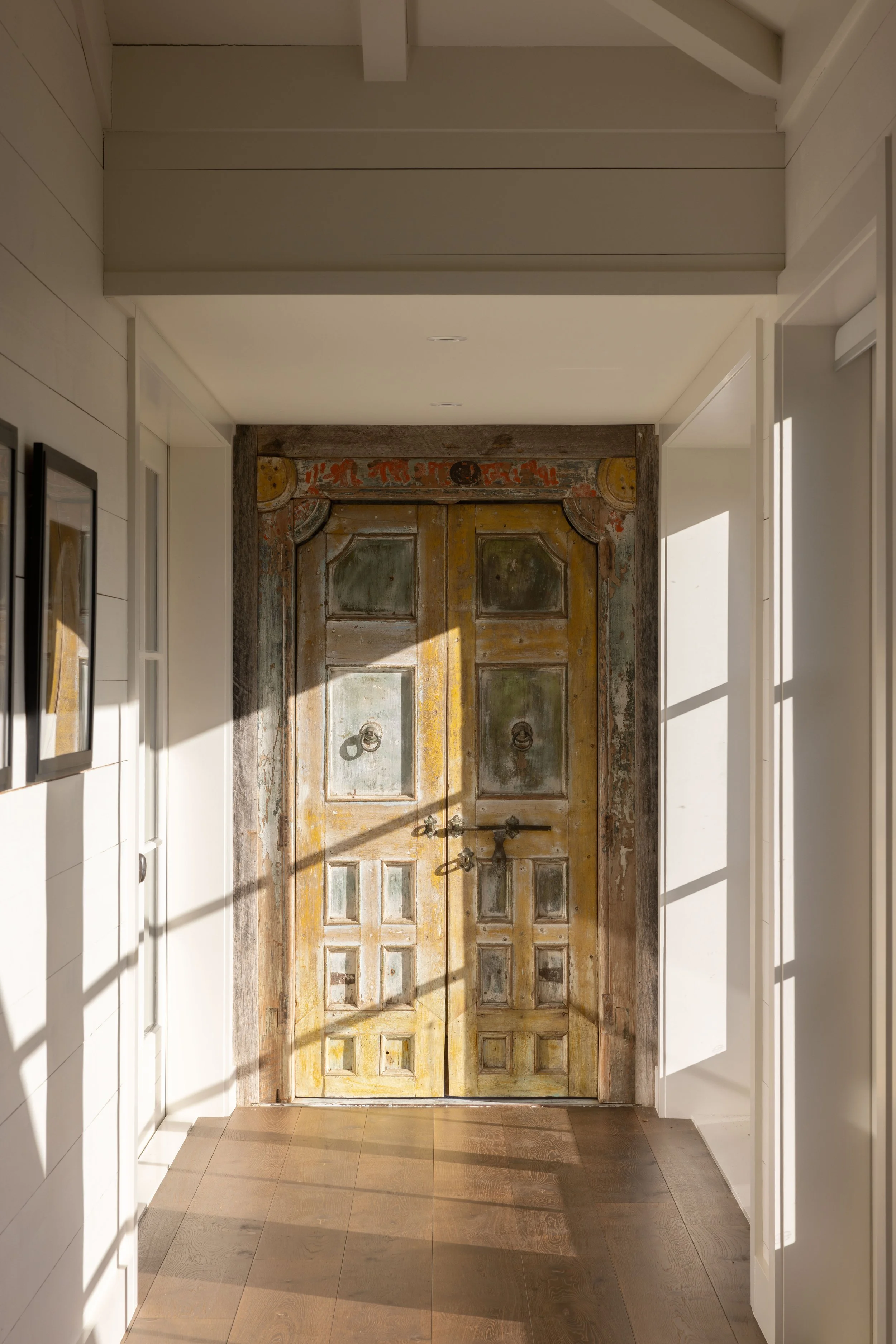 Vintage wooden double door with peeling paint and iron hardware, set in a white modern hallway with sunlight casting shadows.