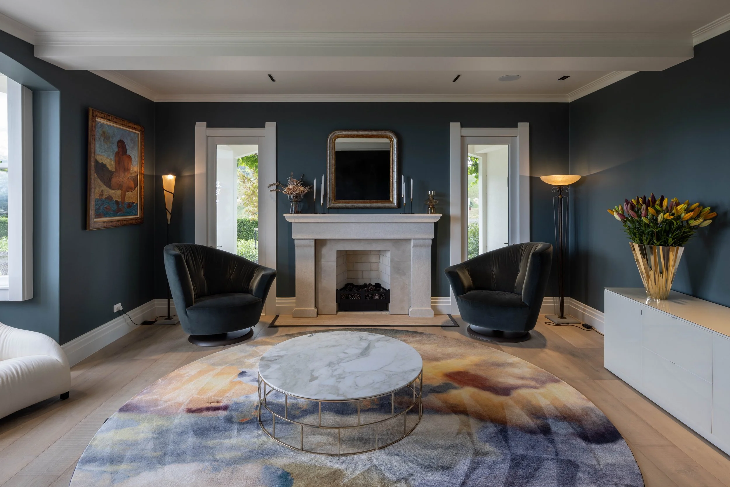 Living room with a fireplace, two black armchairs, a round marble coffee table, and a colorful area rug. Windows on either side of the fireplace display greenery outside. Decor includes artwork, candles, a large mirror, a tall floor lamp, and a white sideboard with a gold vase of flowers.