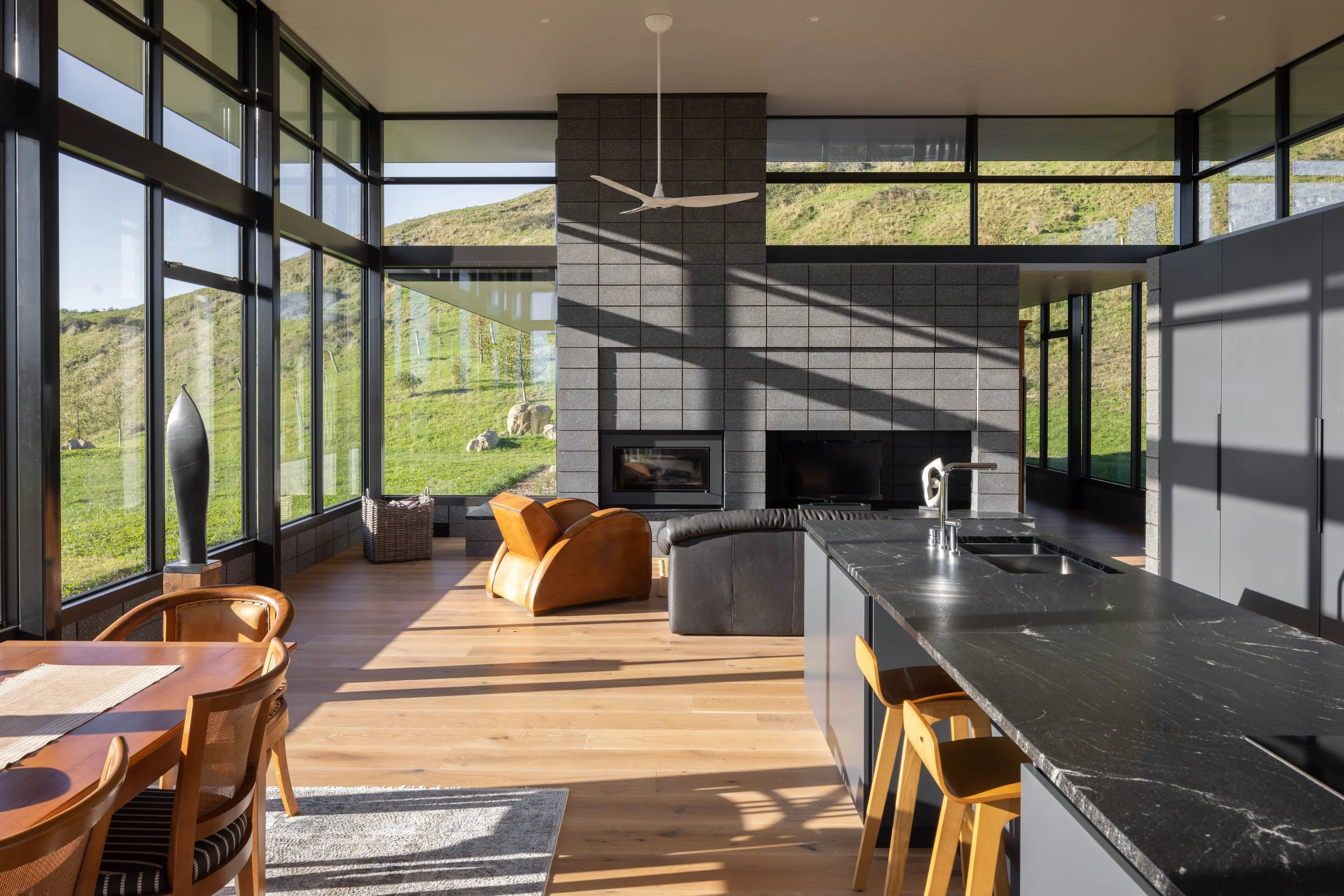 Modern open-concept living space with large floor-to-ceiling windows, a gray brick fireplace, leather and fabric chairs, a black marble kitchen island, and scenic green hills outside.