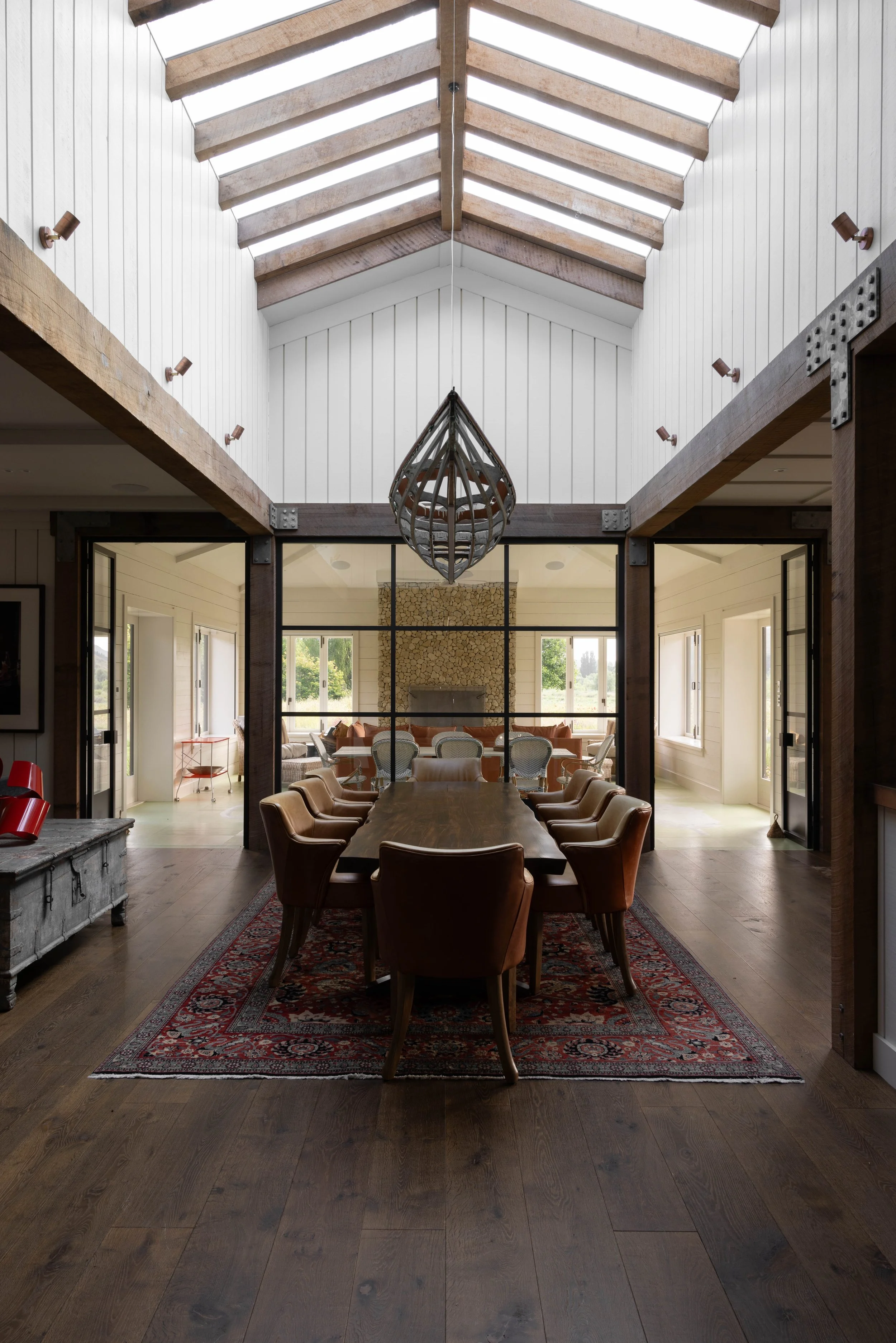 A modern dining room with a high vaulted ceiling, wooden beams, a chandelier, a long wooden table, a warm, rustic design and timber flooring by Terrain Hawke's Bay.