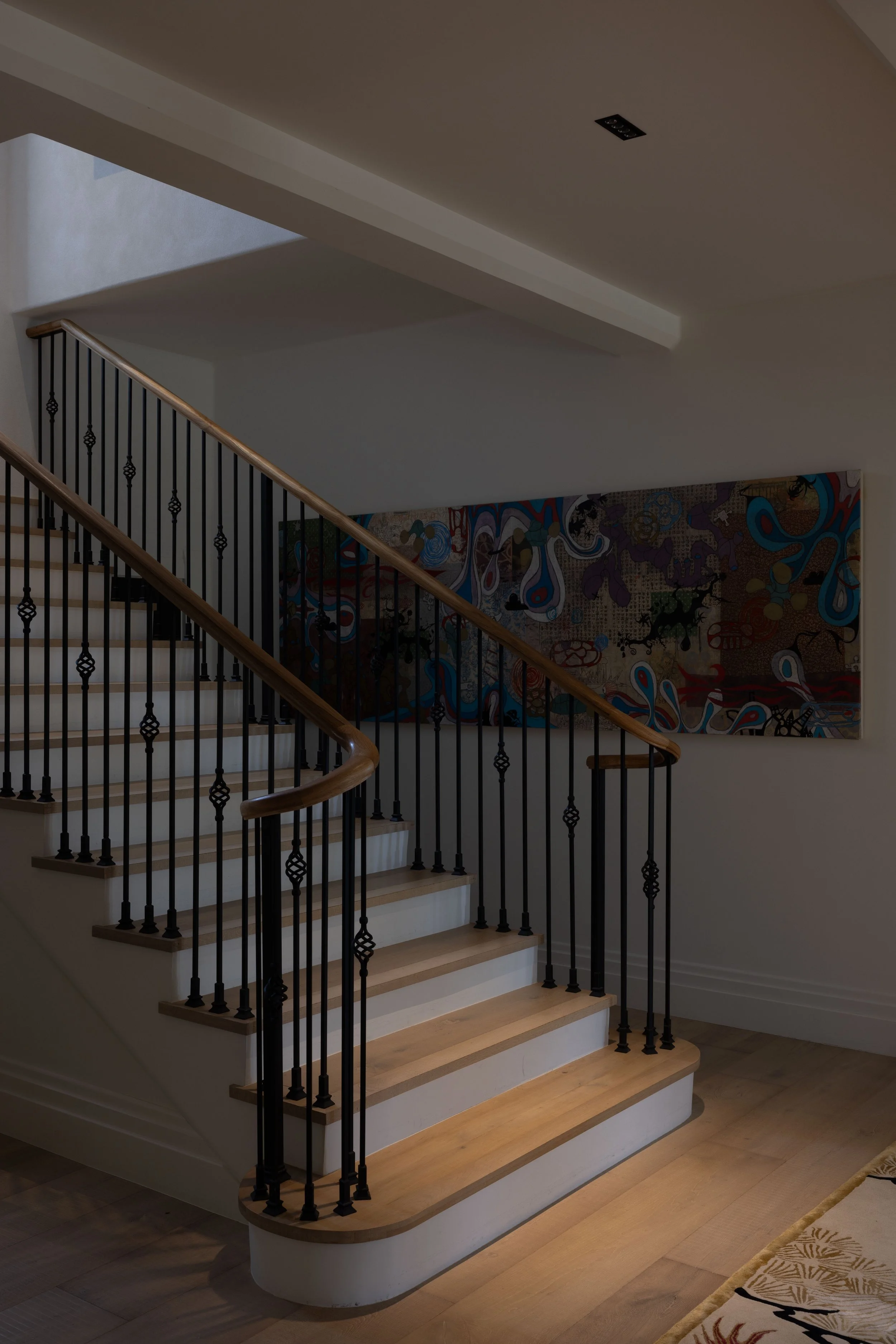 Interior staircase with wooden handrails and black iron balusters, a colorful abstract painting on the wall, and hardwood flooring.
