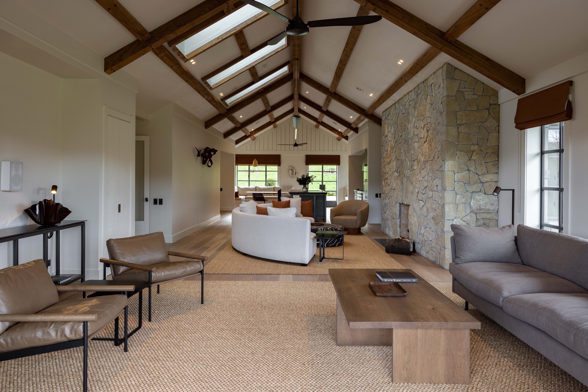 Spacious living room with vaulted ceiling, wooden beams, skylights, stone fireplace, large windows, and modern furniture.