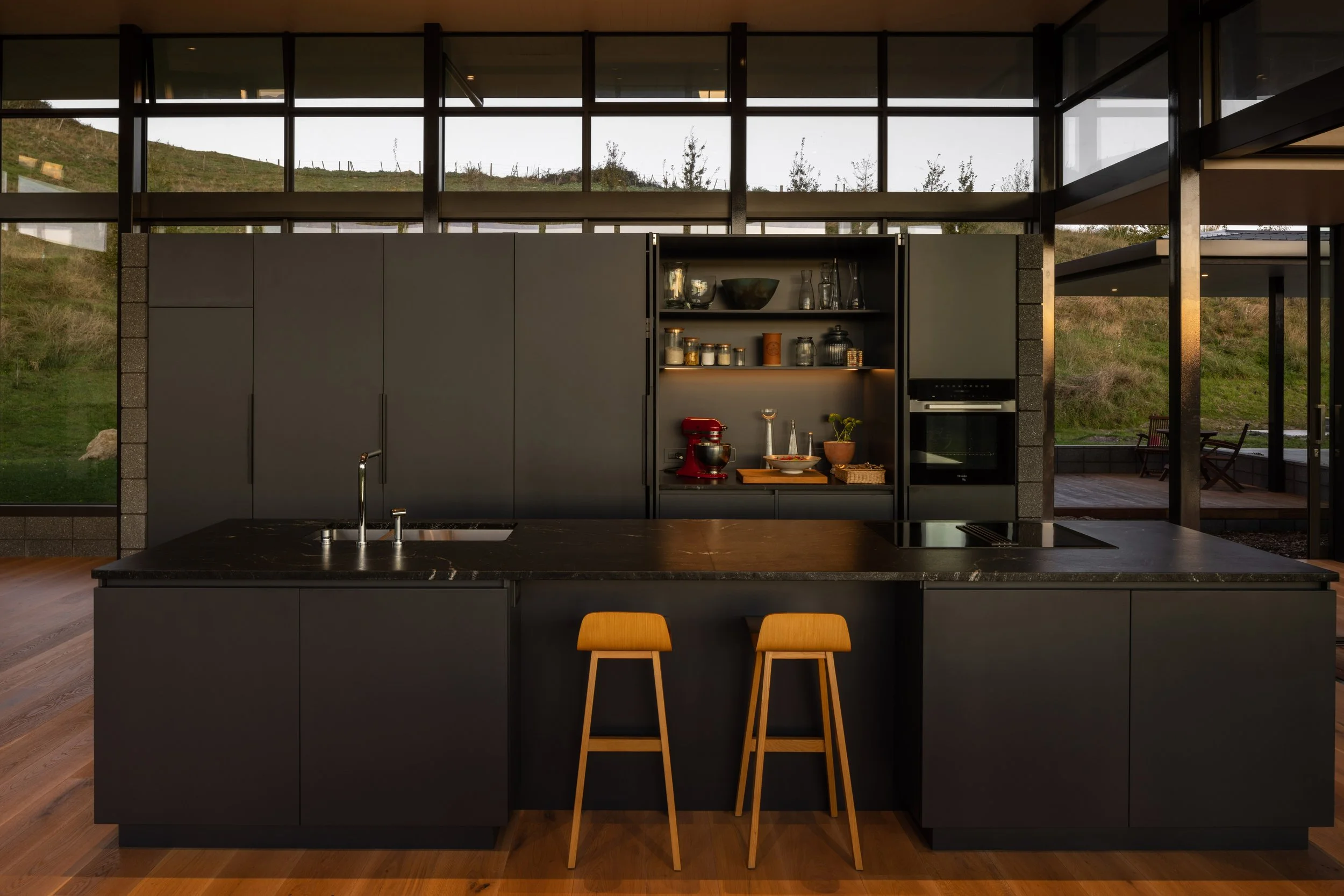 Modern black kitchen with wooden stools, open shelving with kitchenware, large windows showing grassy landscape.