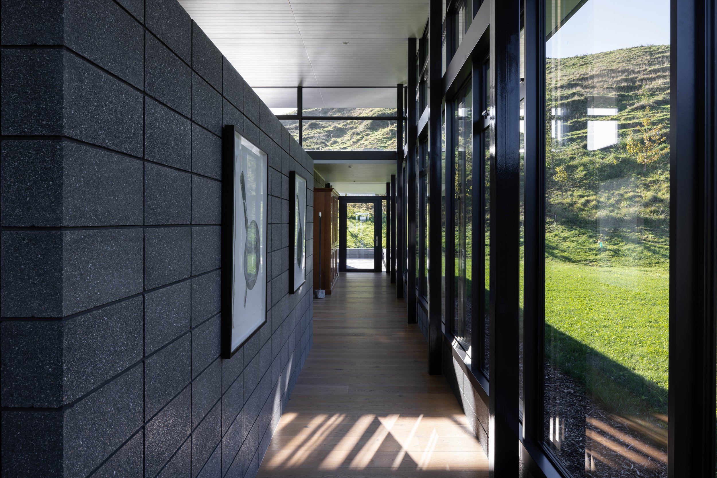 Modern indoor corridor with dark brick wall on the left, large glass windows on the right revealing a green hillside outside, hardwood floor, and artwork on the wall.