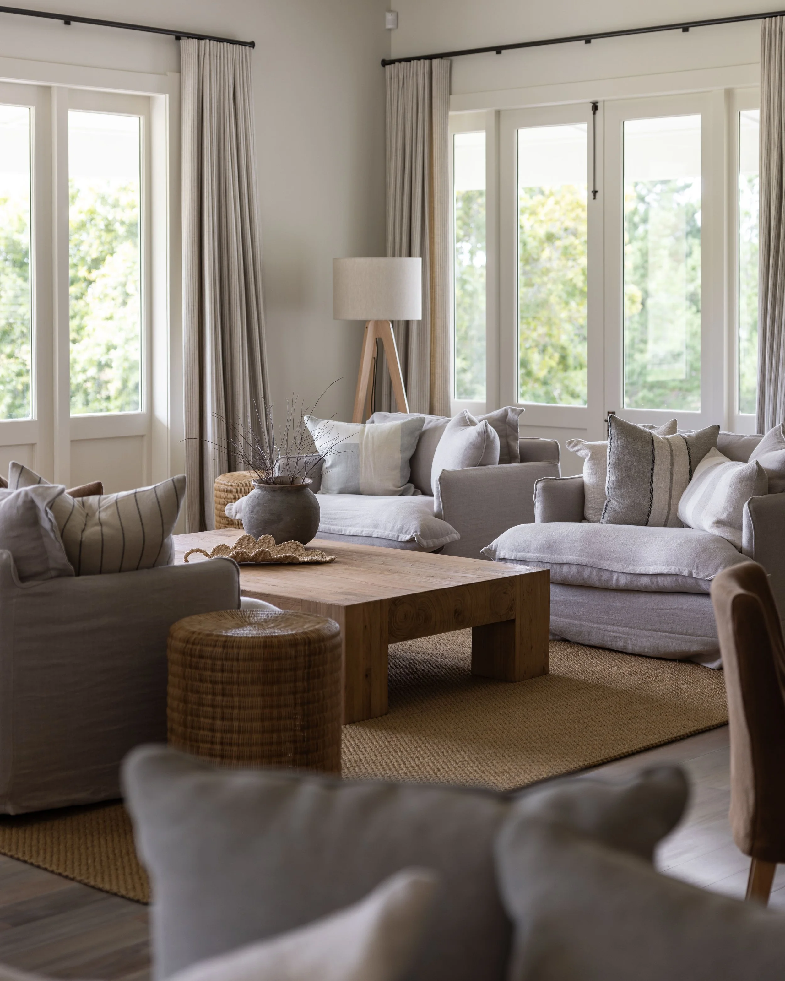 A cozy living room with large windows, beige sofas with pillows, a wooden coffee table, a wicker ottoman, a floor lamp, and neutral curtains.