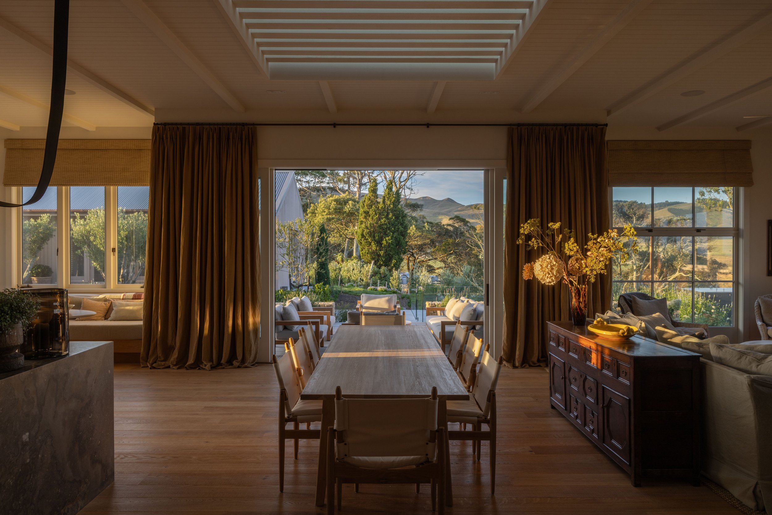 Interior of a dining room with a large wooden table, opening to an outdoor patio with seating. Beautiful timber flooring by Terrain, Hawke's Bay.