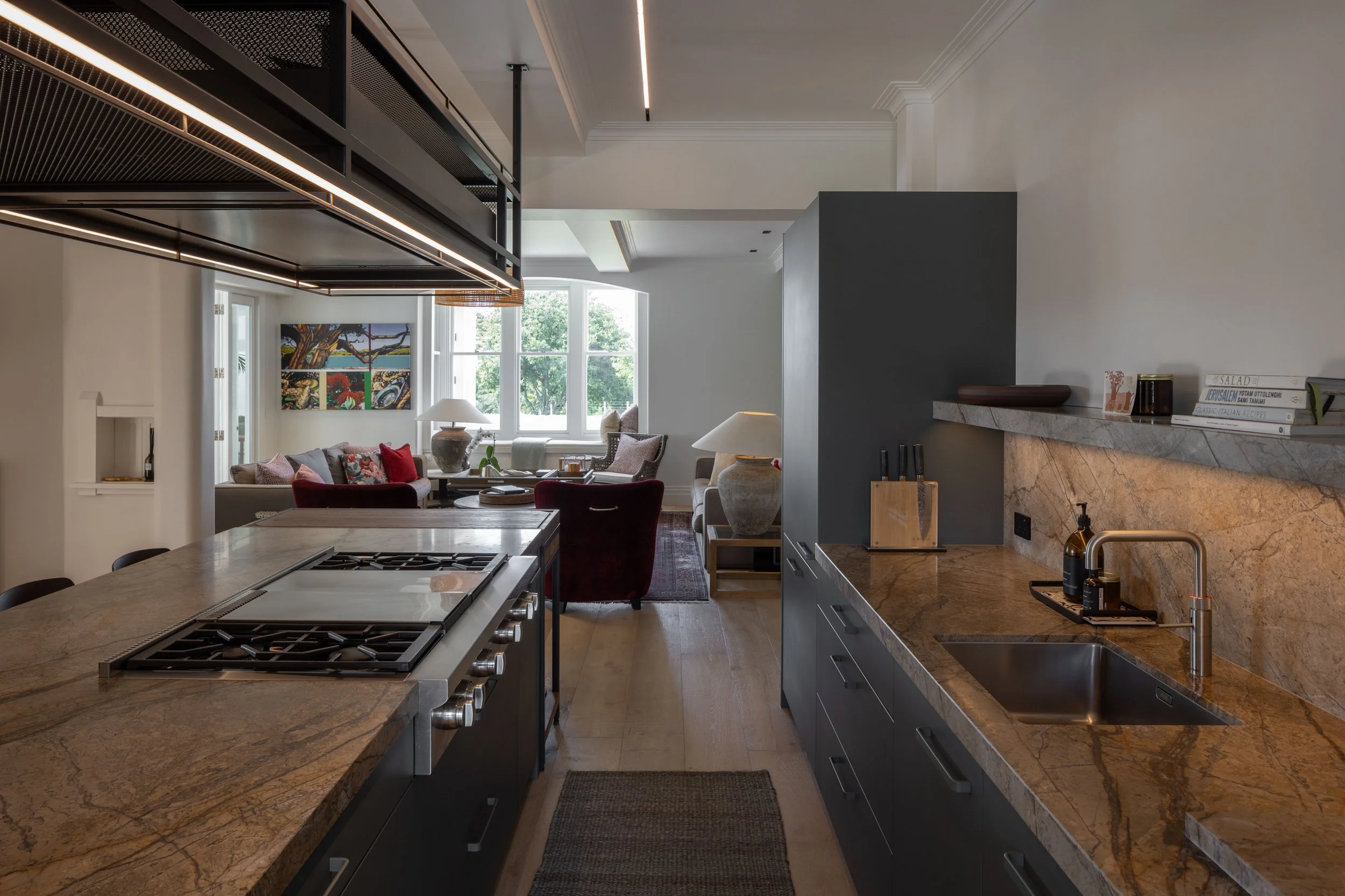 Modern kitchen with marble countertops, stove, sink, gray cabinets, and connected living room with large window, artwork, and seating.
