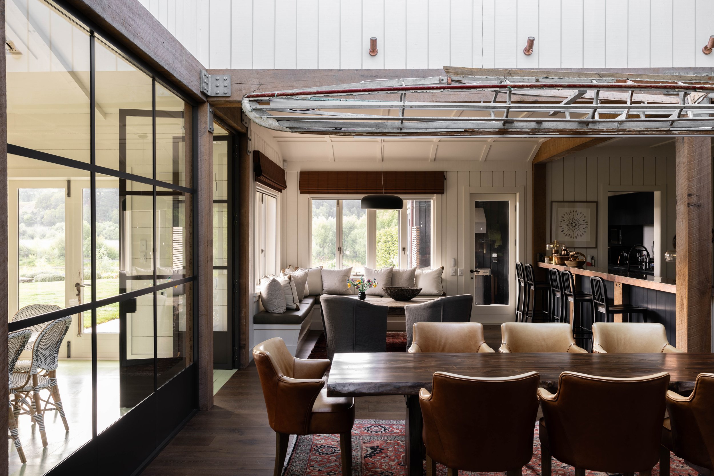 Interior view of a dining and living area in a house with large windows providing a view of greenery outside. The dining table has brown leather chairs, and the living area features a built-in window seat with pillows, a dark armchair, and a small ta