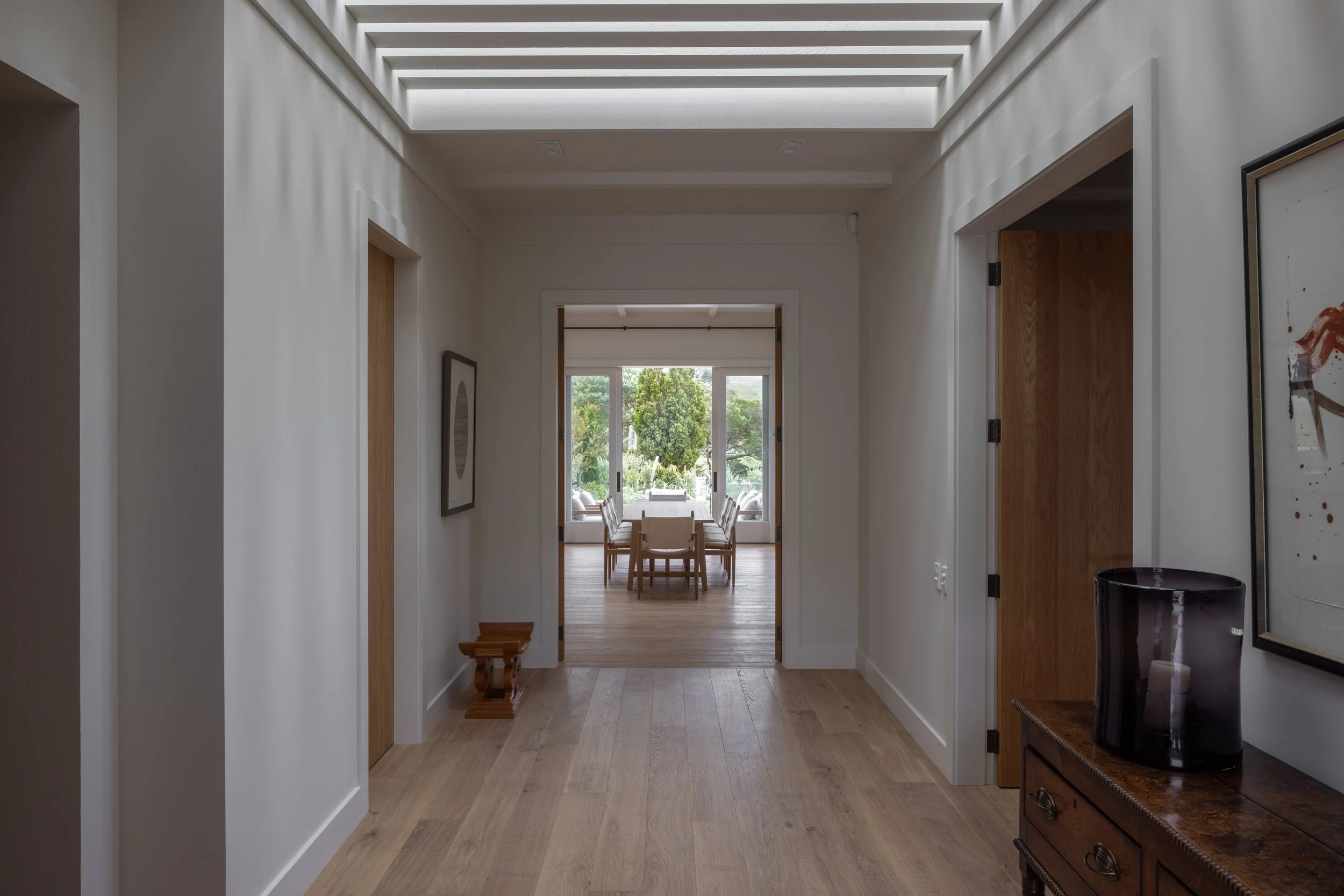 View of a hallway leading into a dining area with large windows and outdoor greenery.