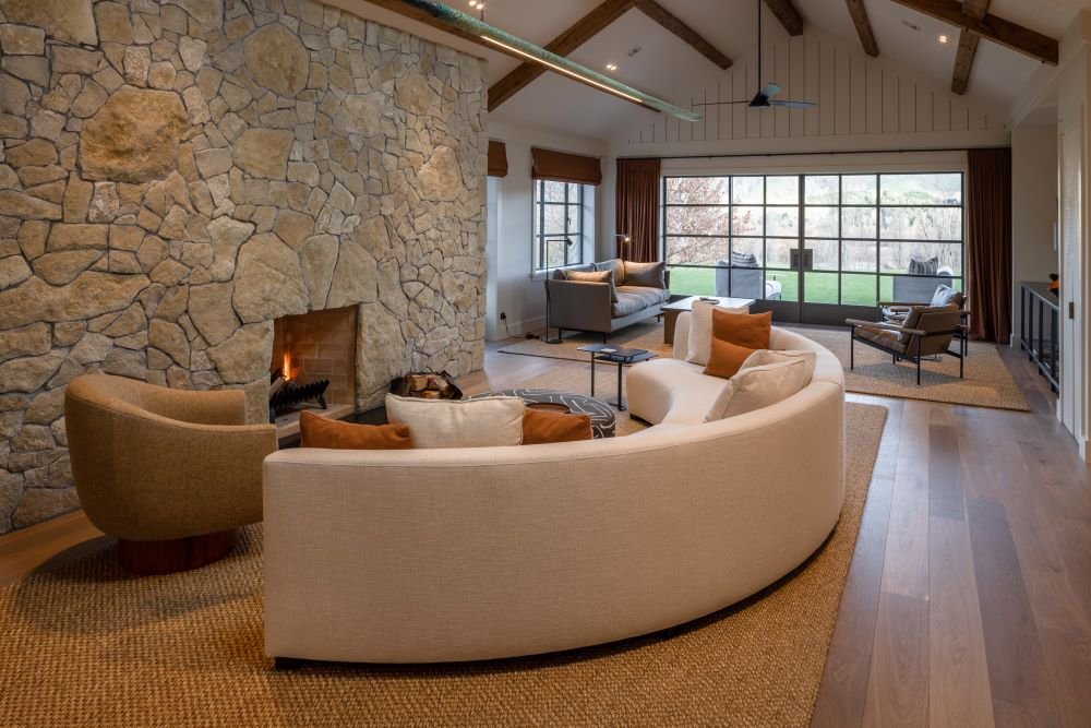 Living room with a large stone fireplace, curved sofa, armchair, and large windows showing a backyard.