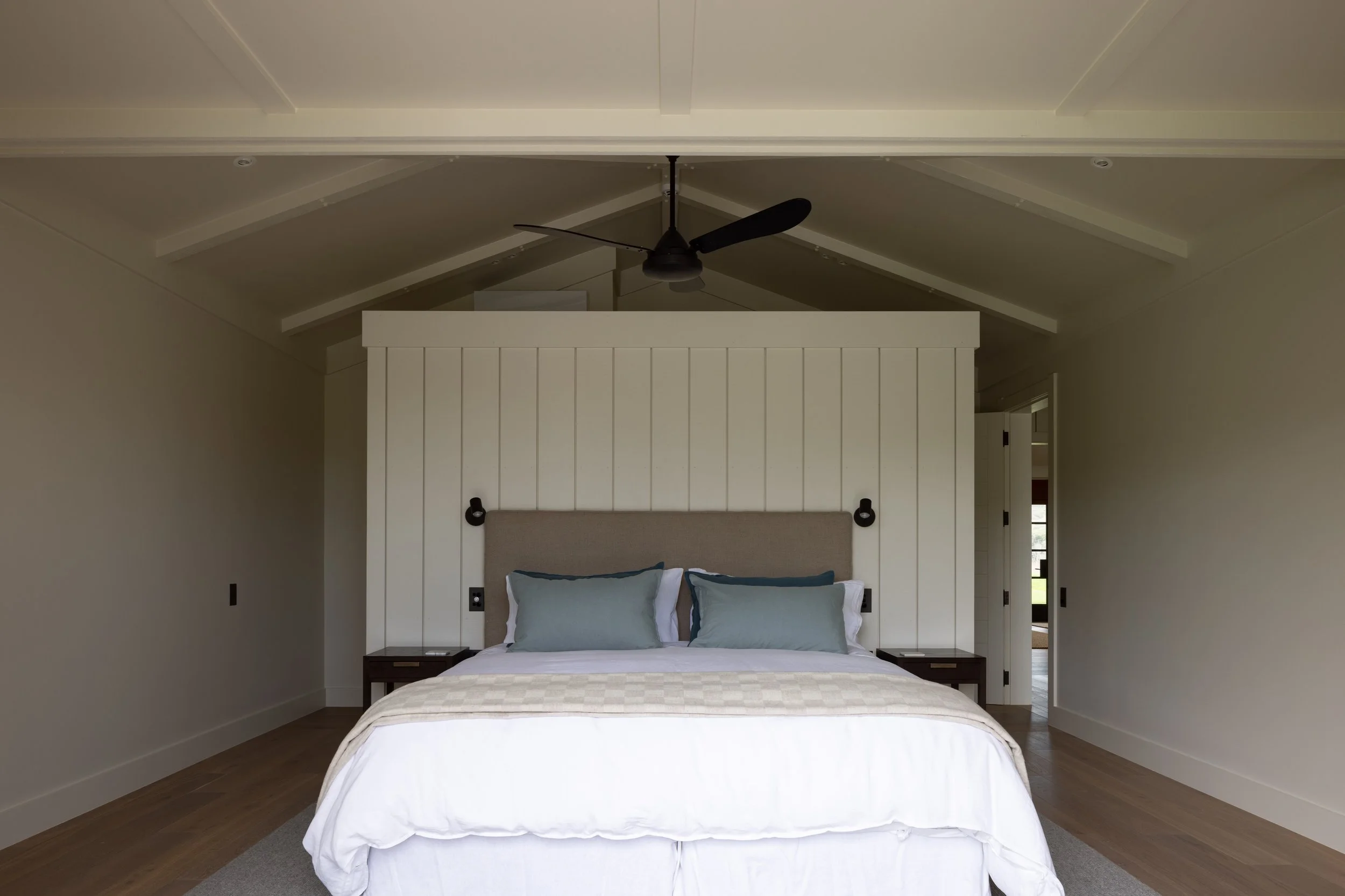 A minimalist bedroom with a large bed, gray headboard, two blue pillows, and white bedding, flanked by two small dark wooden nightstands, in front of a white shiplap accent wall, with ceiling fan above, in a bright room with light-colored walls and h