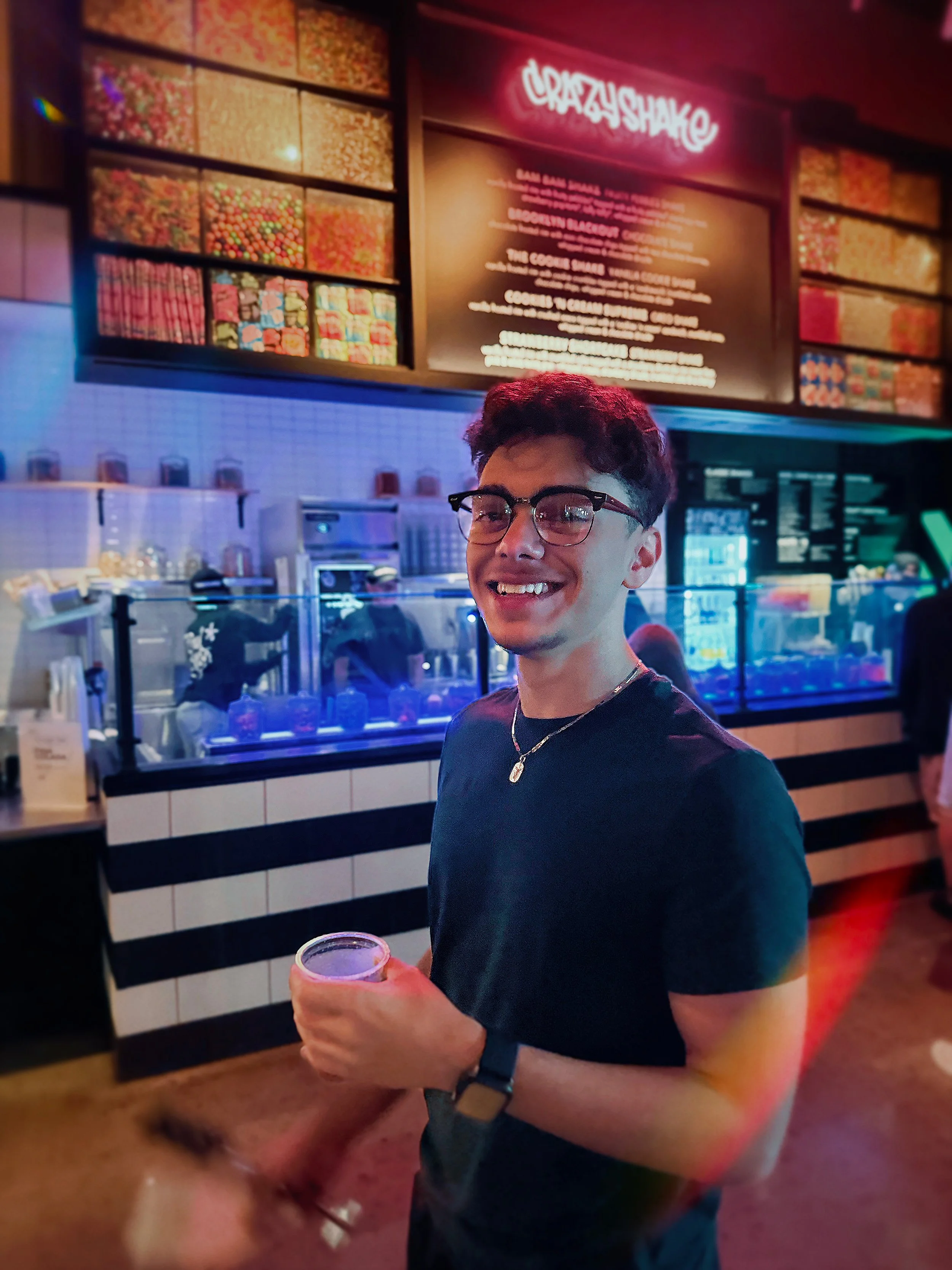 Photo of Kevin Salama. A young man smiling with glasses, holding a drink, standing inside a colorful candy store called 'Crazy Shake' with a menu board behind him.