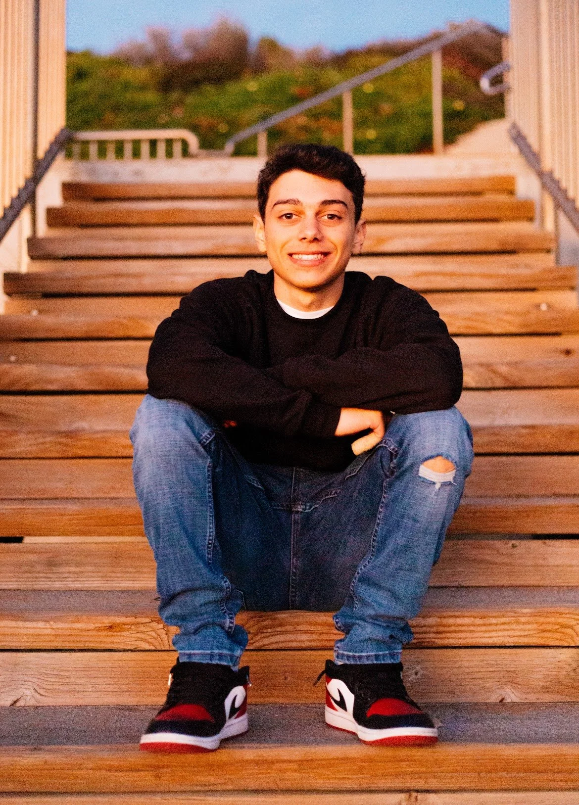 Photo of Kevin Salama. A young man with short dark hair smiling, sitting cross-legged on wooden outdoor stairs, wearing a black sweater, ripped jeans, and black and red sneakers, with a green hill and railing in the background.