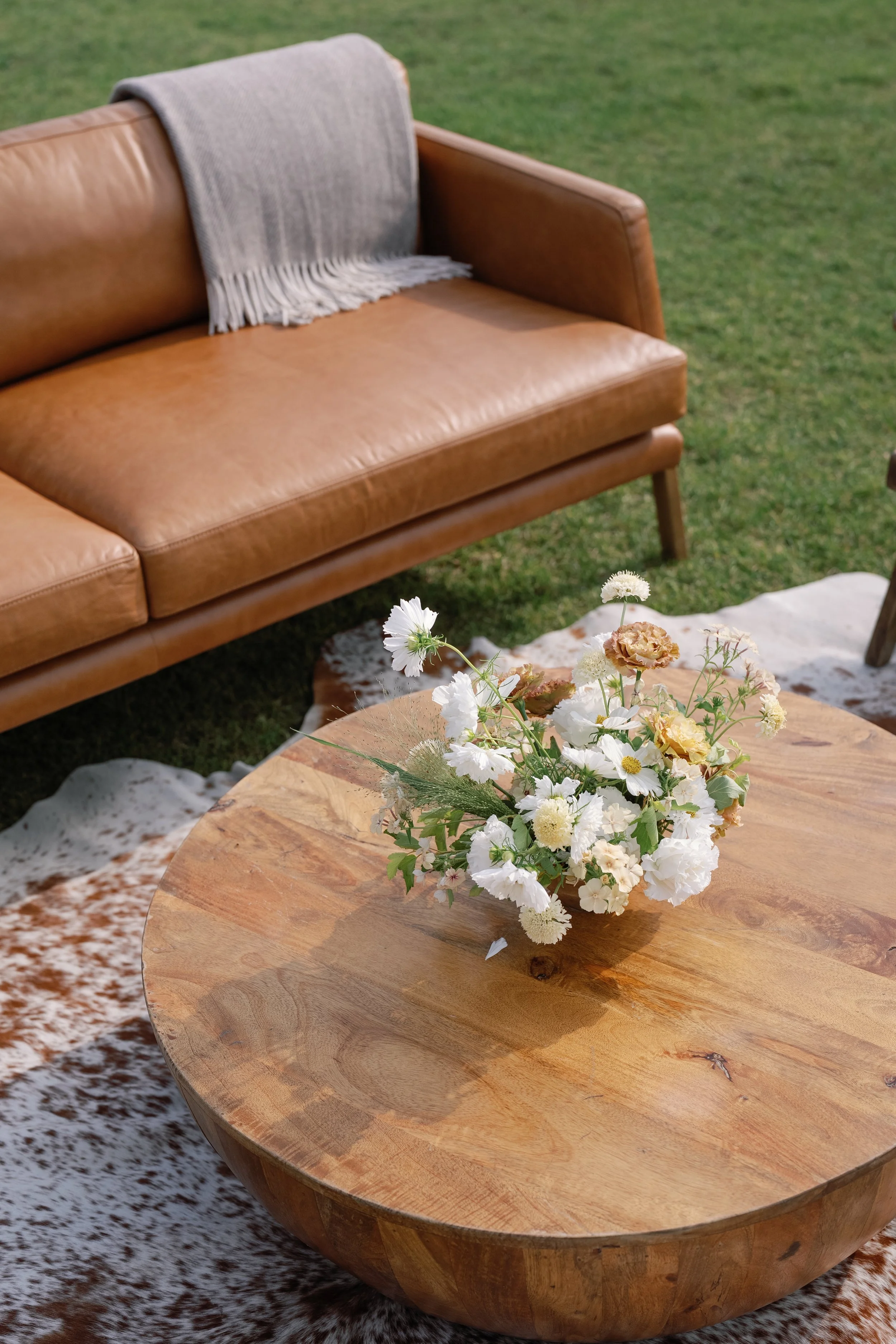 A round wooden table with a bouquet of white and beige flowers. In the background is a tan leather sofa with a gray throw blanket, set outdoors on a grassy area.