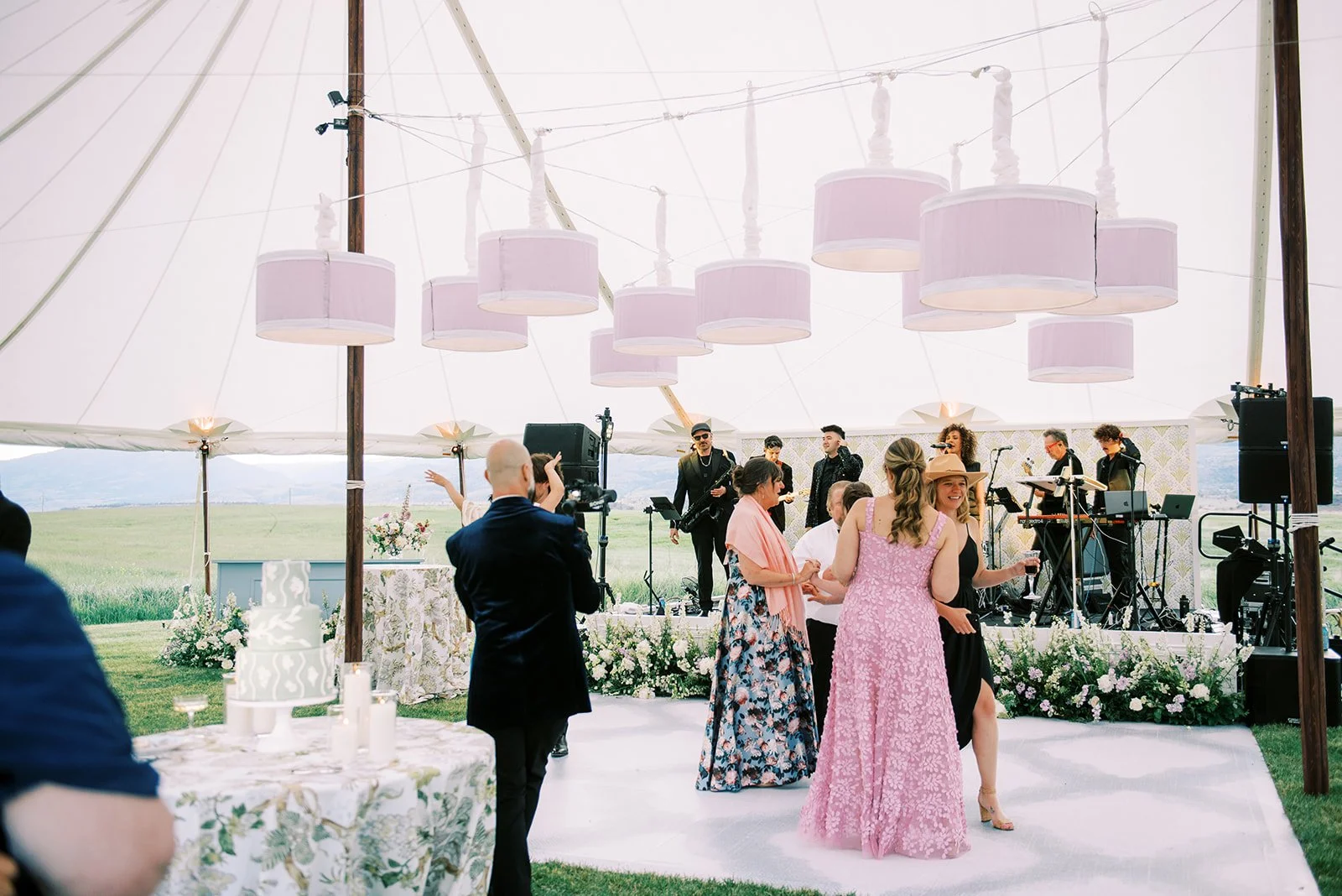 People dancing and socializing at an outdoor wedding reception under a tent with large hanging pink lamps and a live band playing in the background.
