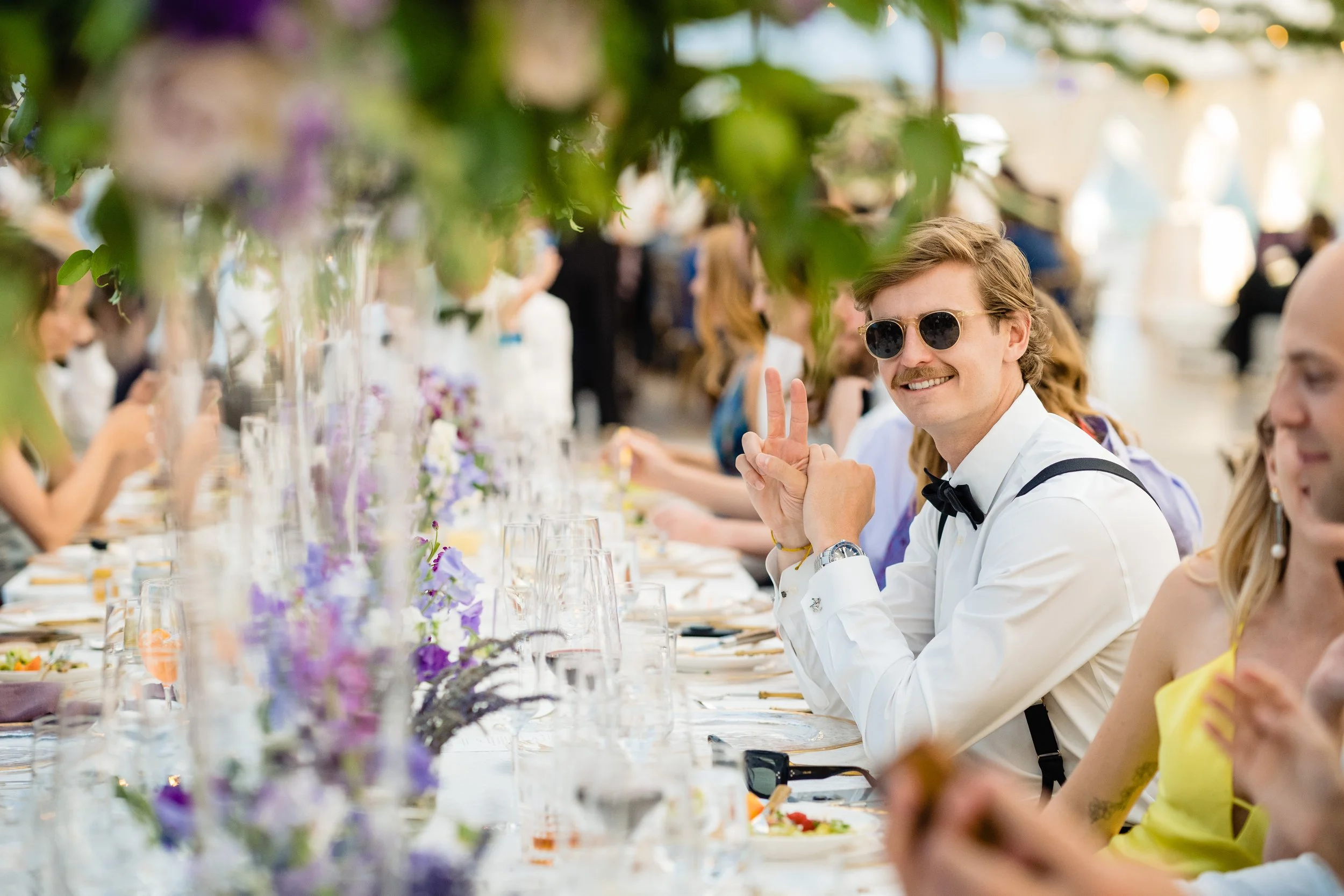 A man with curly blond hair, wearing sunglasses and a white shirt, making a peace sign at a long outdoor restaurant table. The table is decorated with purple and white flowers, and there are other people dining and socializing in the background.