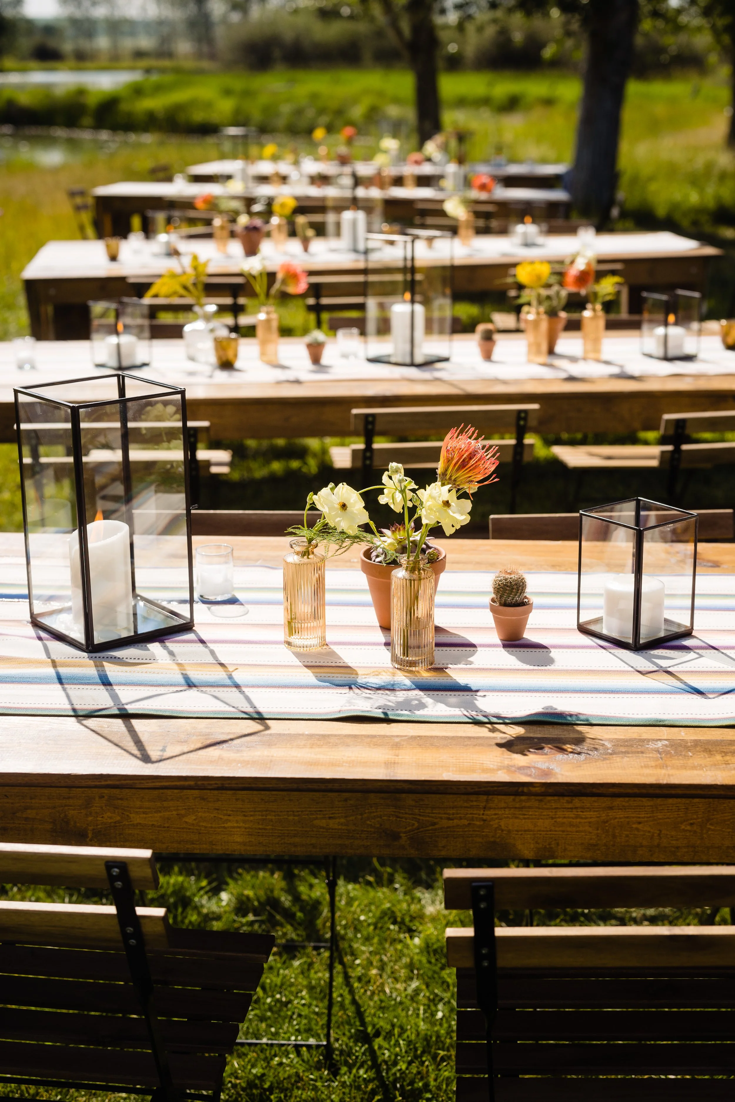Outdoor dining setup with wooden tables decorated with flowers and candles, set in a grassy area next to a pond with trees in the background.