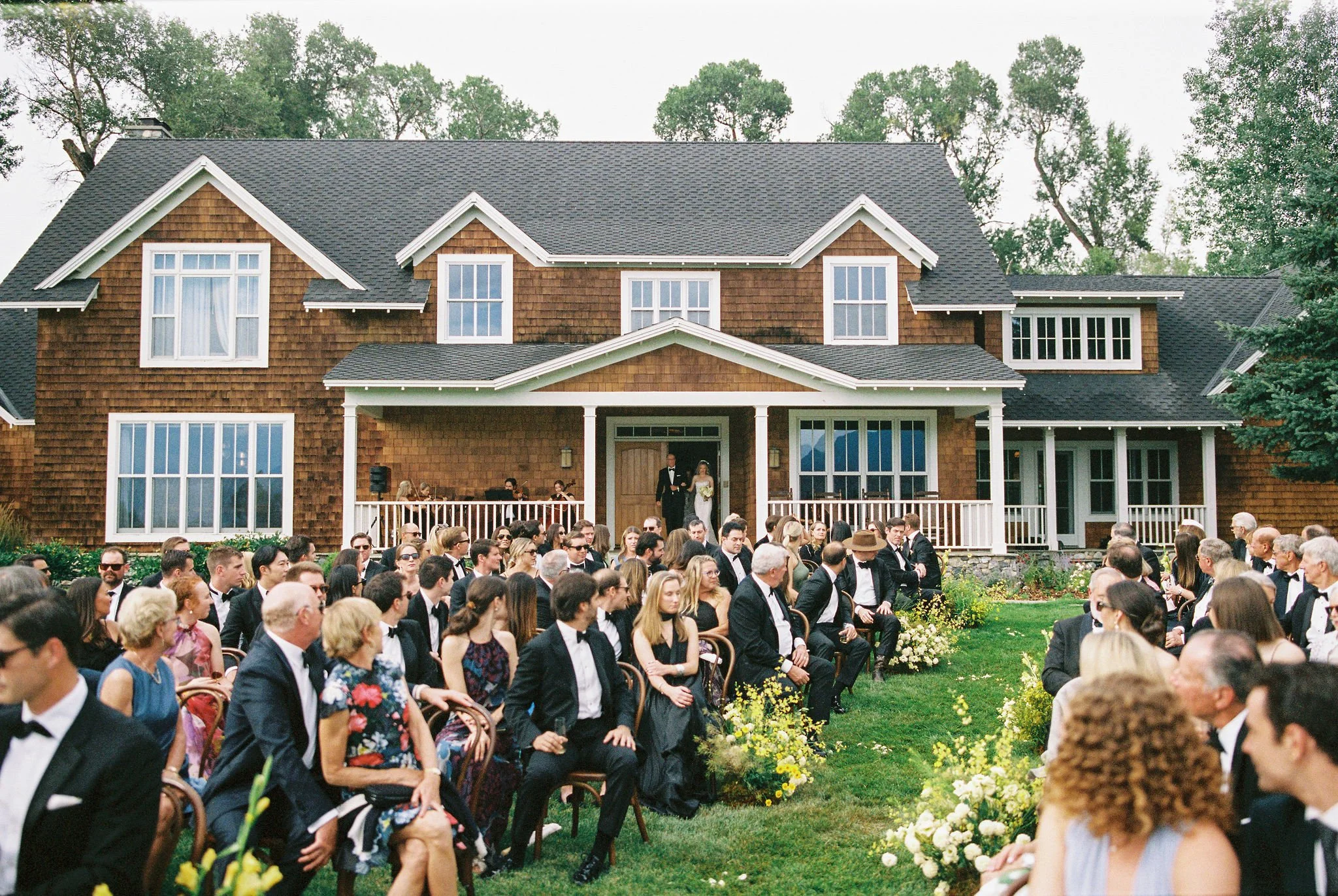 People gathered at an outdoor wedding ceremony in front of a large brown house with a porch, surrounded by trees.