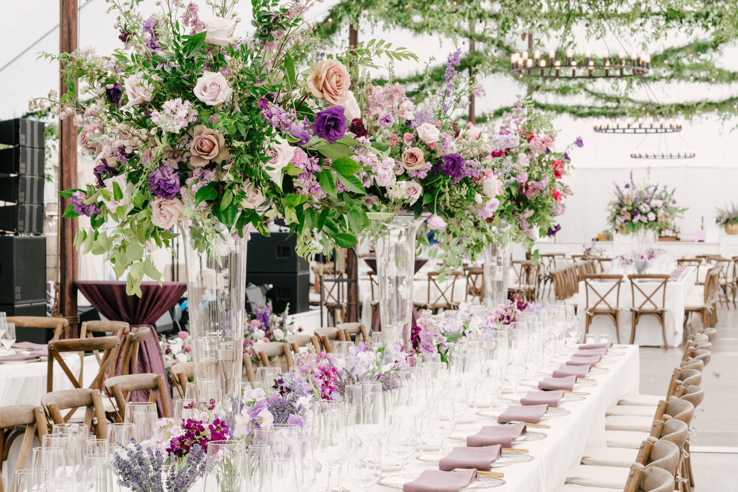 Elegant wedding reception setup in a bright, decorated tent with long tables, purple and pink floral centerpieces, and hanging chandeliers.