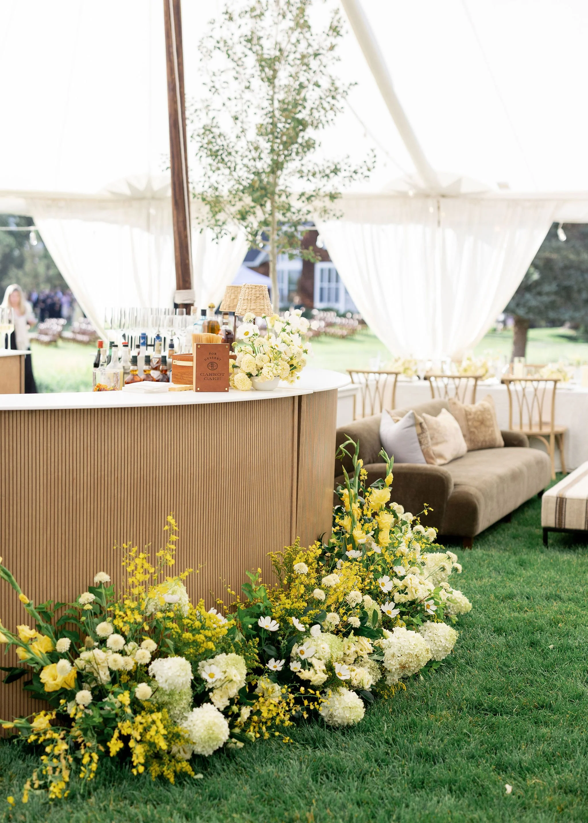 White and yellow floral arrangement styled in front of a wooden bar under a sailcloth tent at a Montana wedding, by Wild Blume