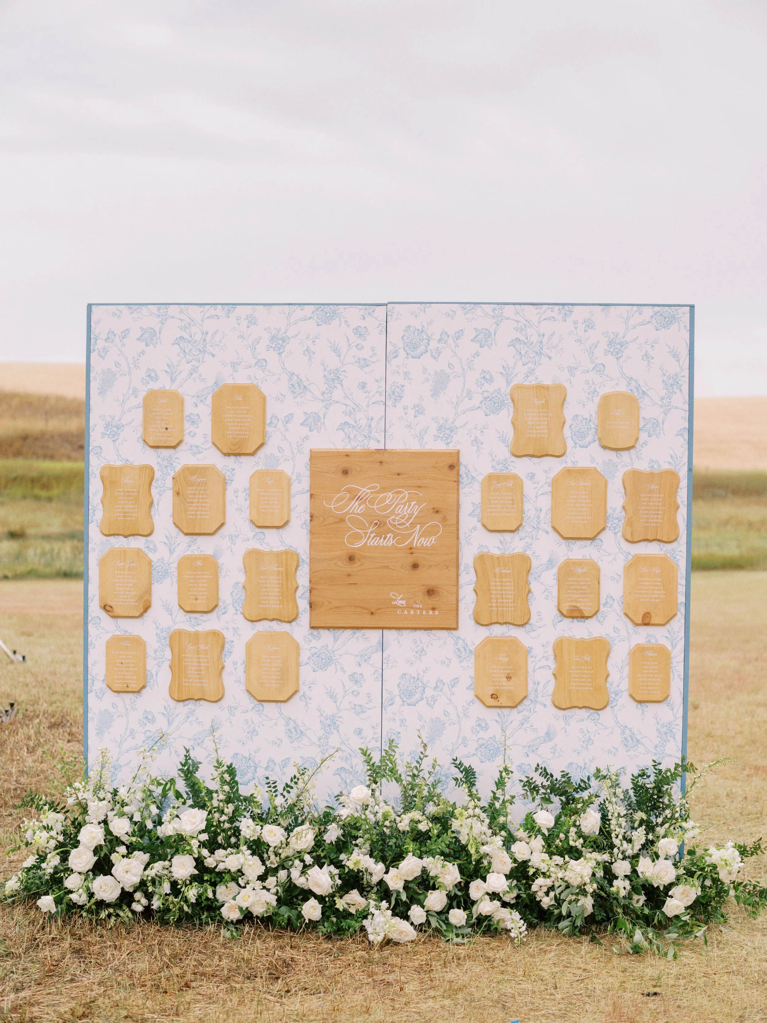 A wedding seating chart display with a pink floral backdrop, wooden name plaques, and white floral arrangements at the bottom, set outdoors on a grassy field.
