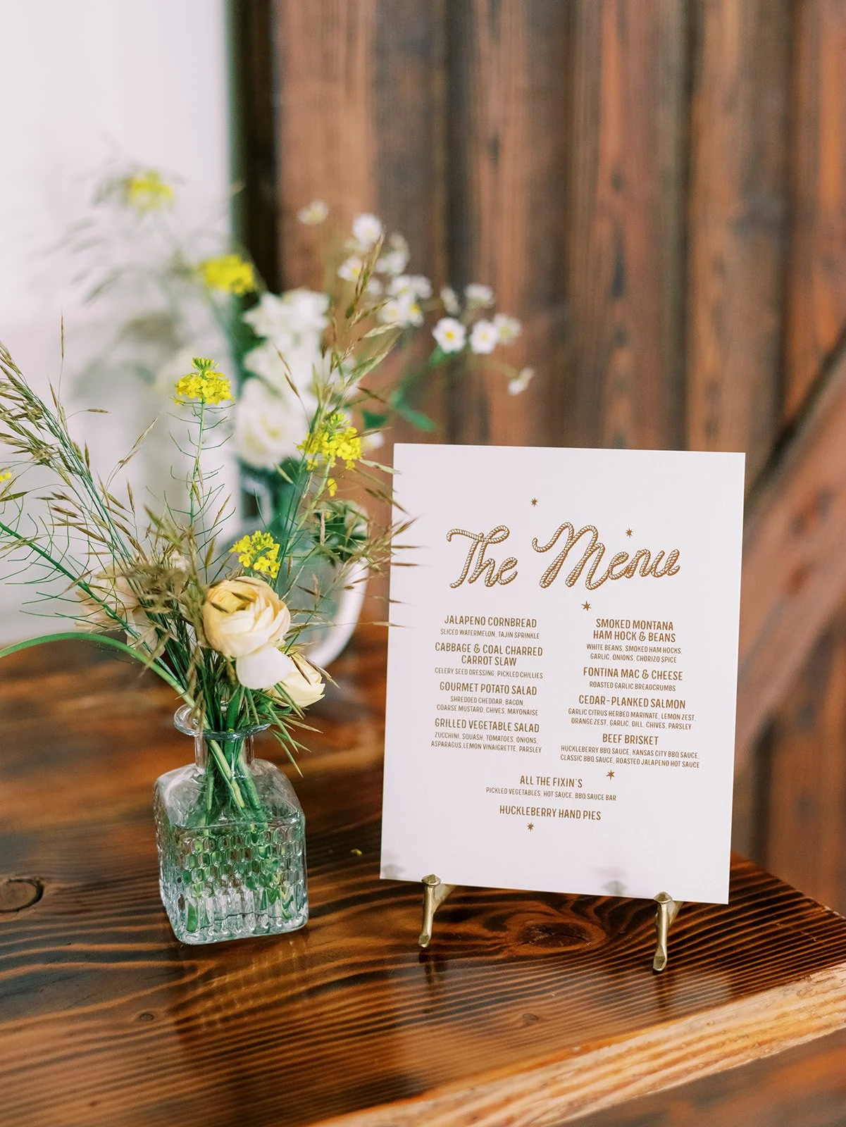 A menu card on a small stand and a vase with flowers on a wooden table, with a blurred background of a wooden wall.