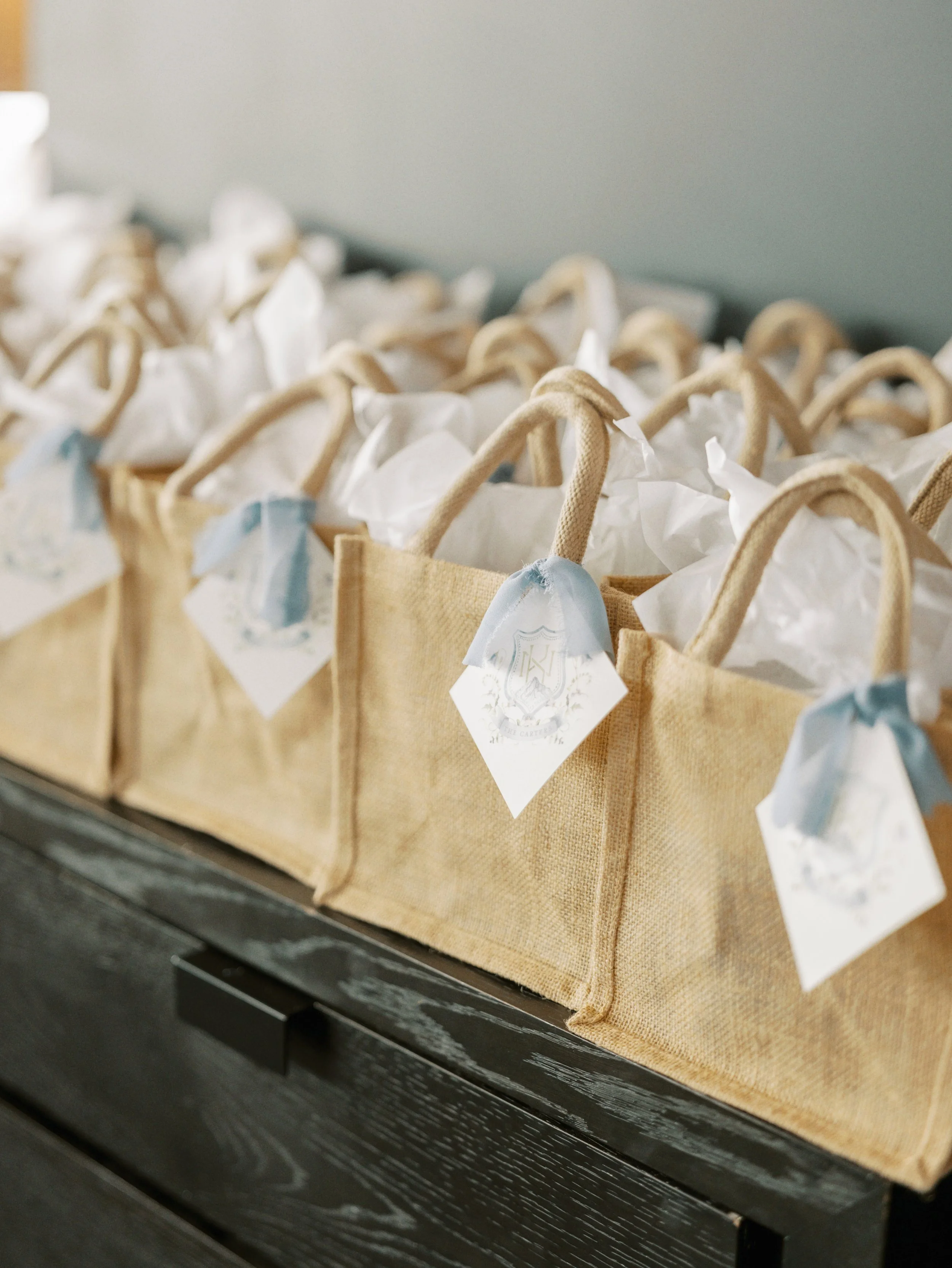 Multiple beige paper bags with twisted paper handles, each with a decorative white and blue tag attached, arranged on a dark wooden surface.