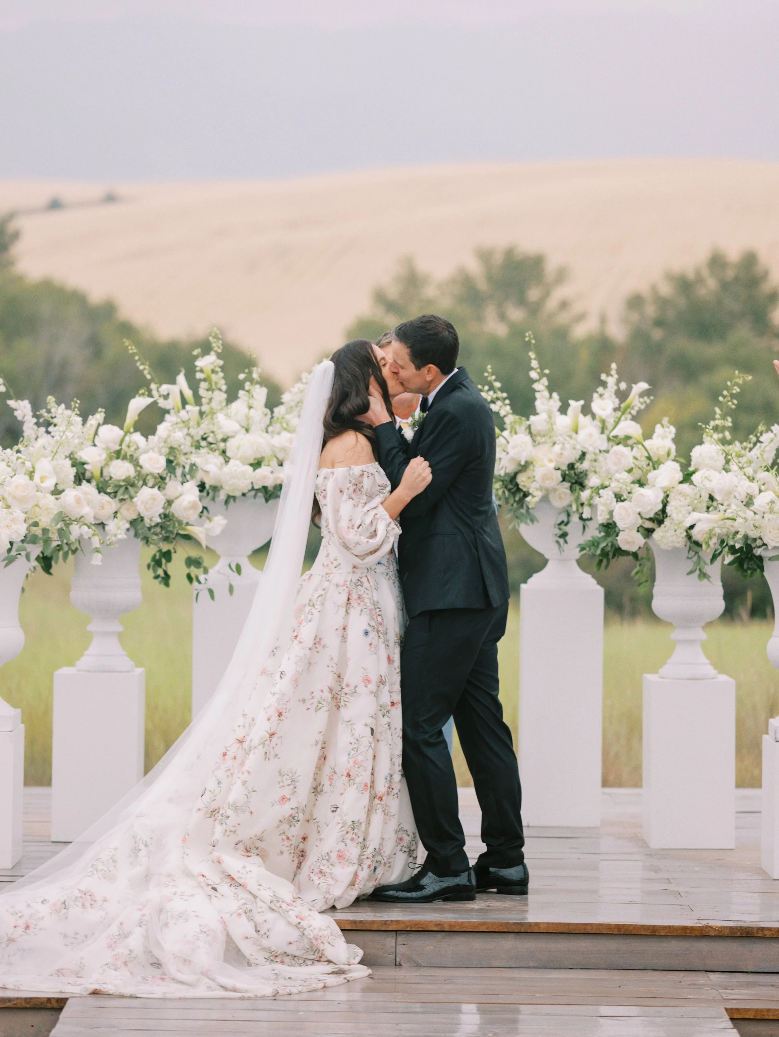 A bride and groom sharing a kiss during their wedding ceremony outdoors, surrounded by white floral arrangements and white urns on pedestals, with a landscape of rolling hills and trees in the background.