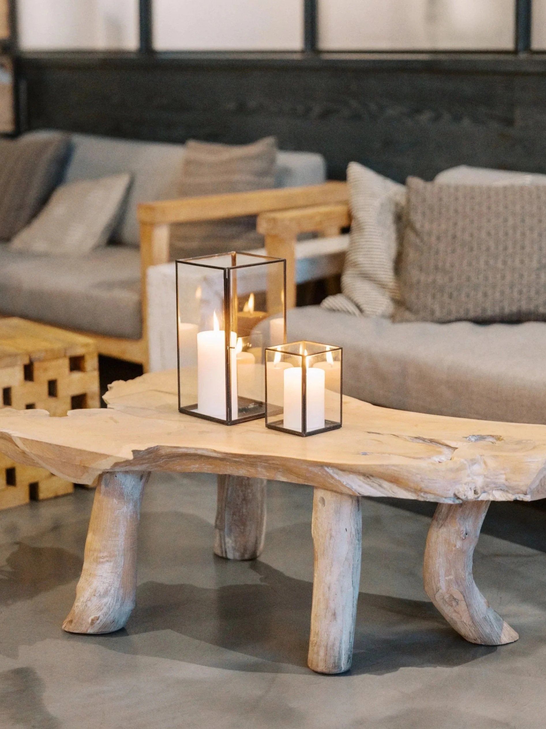Rustic wooden table with a glass lantern containing candles, set in a cozy living space with sofa pillows and a dark wall with windows in the background.