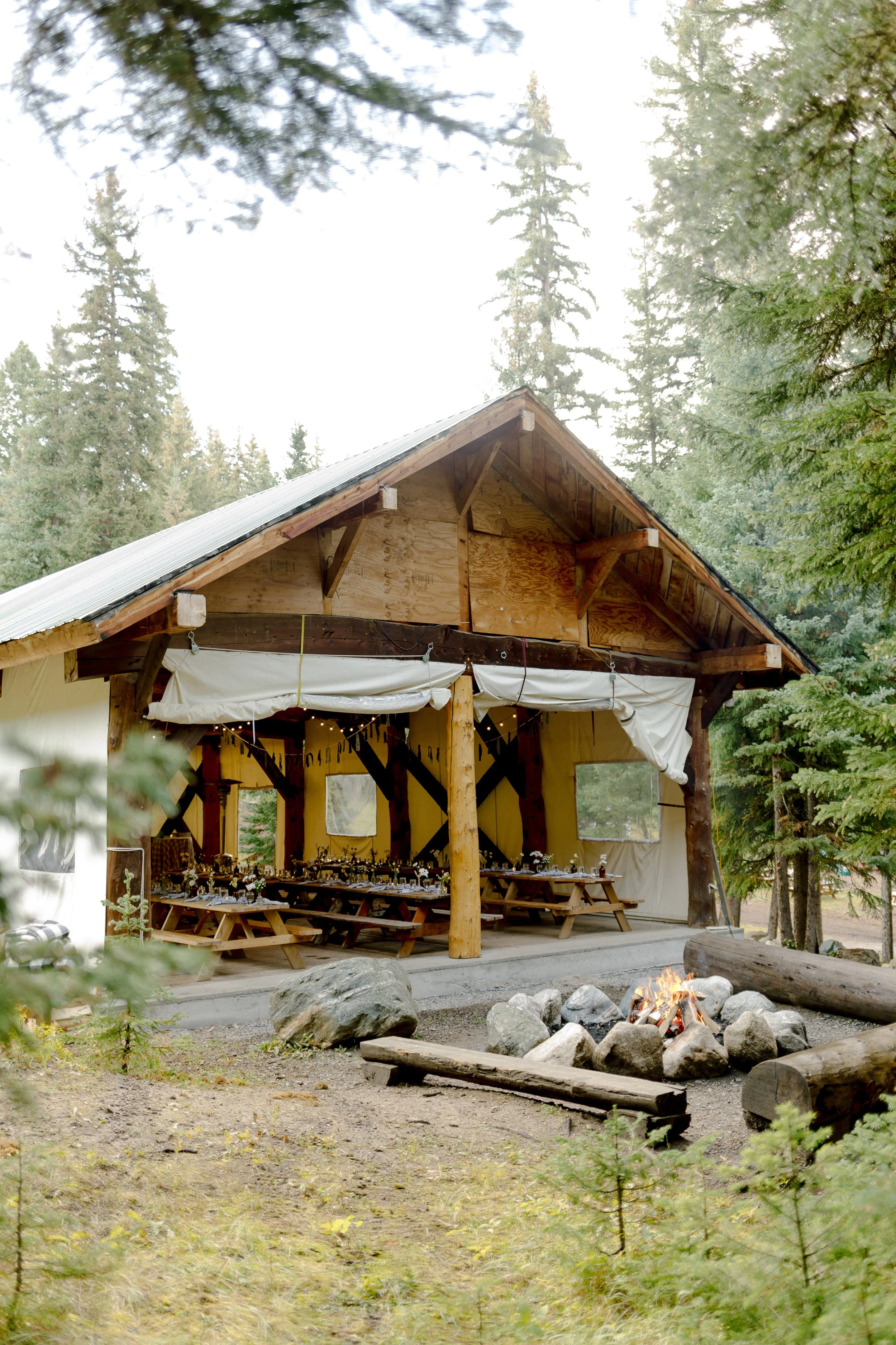 Open-air wooden shelter with string lights, set in a forest with a campfire and logs in the foreground.