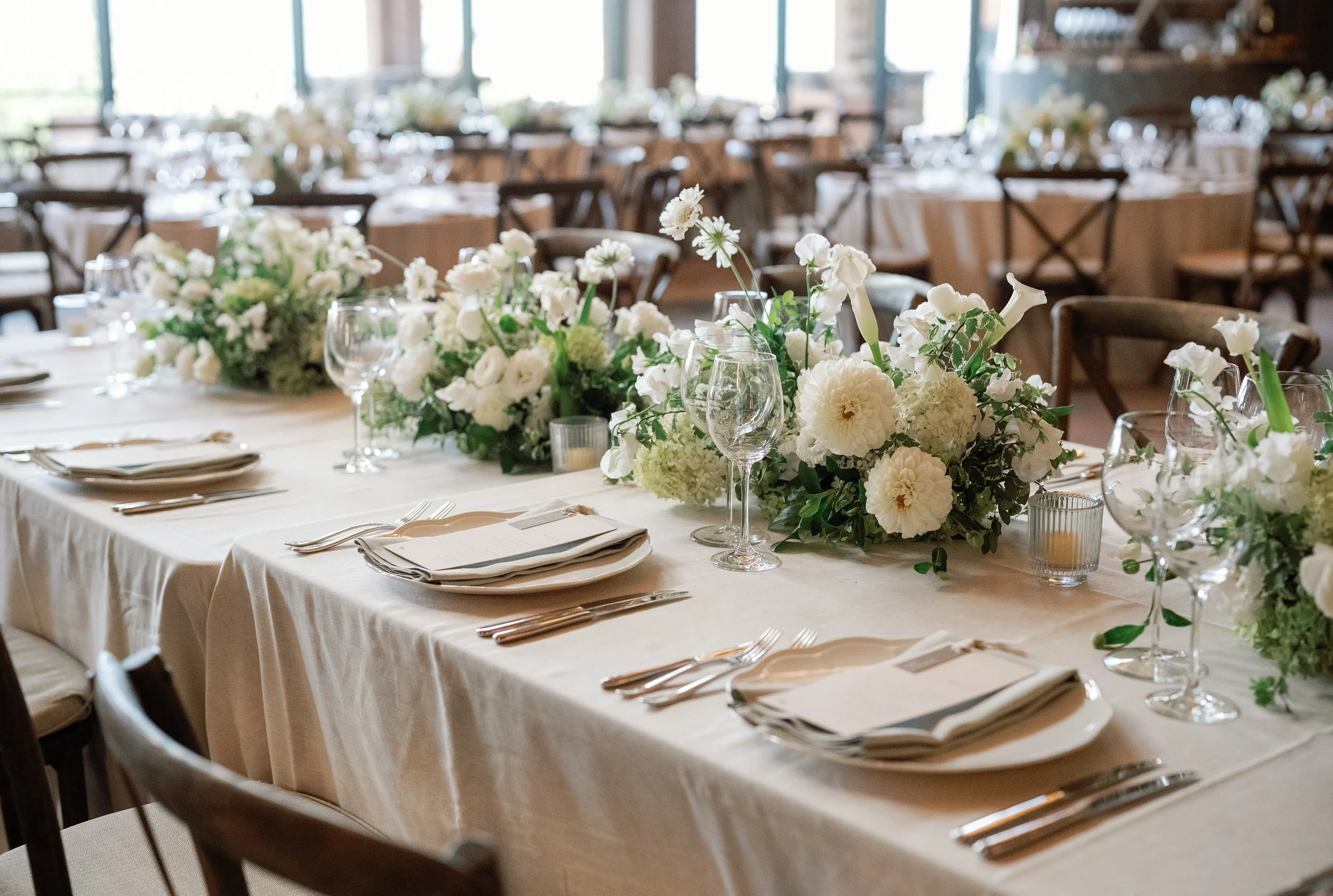A banquet table decorated with white floral centerpieces, wine glasses, wrapped silverware, and table settings in a bright, rustic hall with large windows.