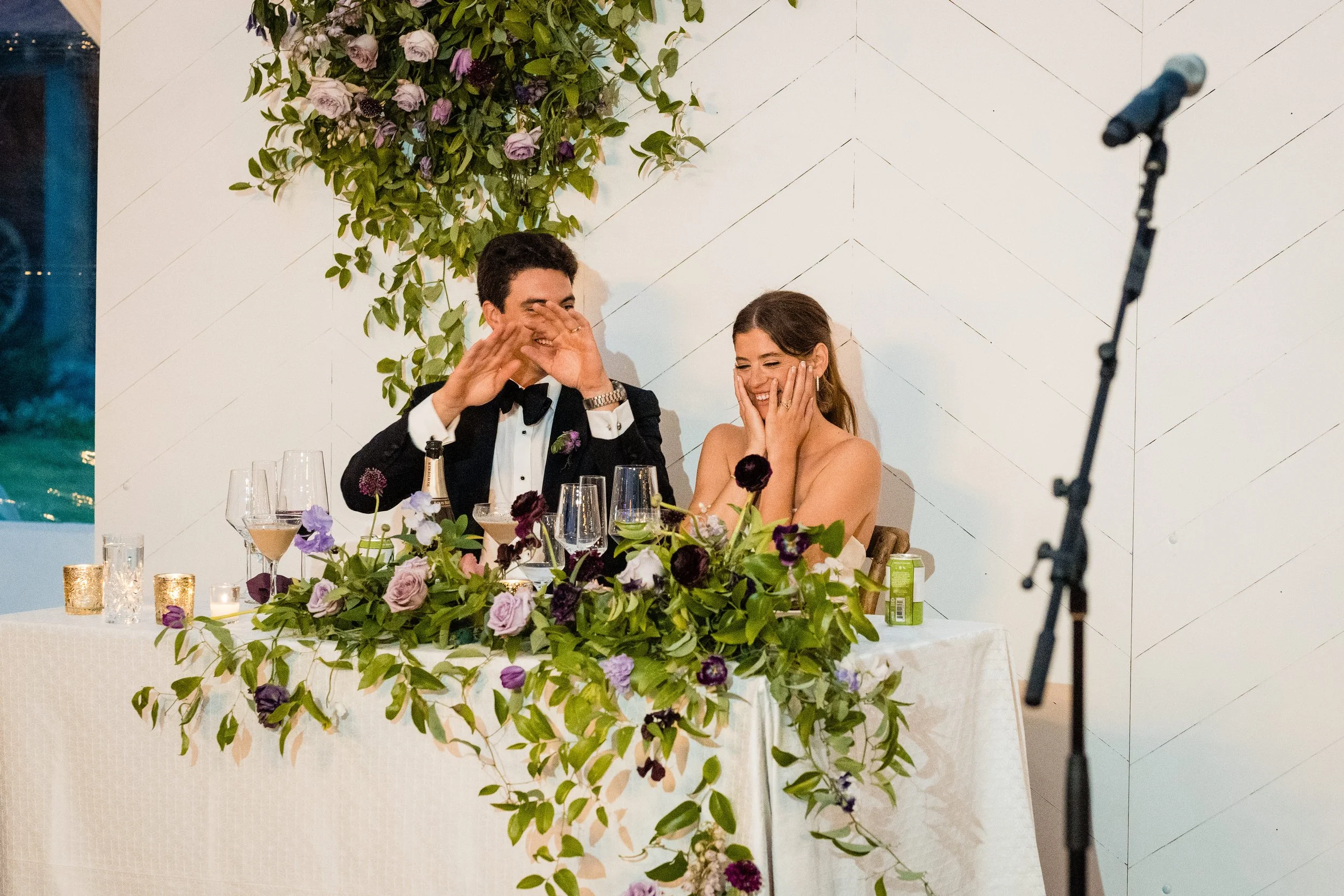 A couple dressed in formal attire sitting at a decorated table with flowers, laughing and covering their faces. There is a microphone on a stand in the foreground.