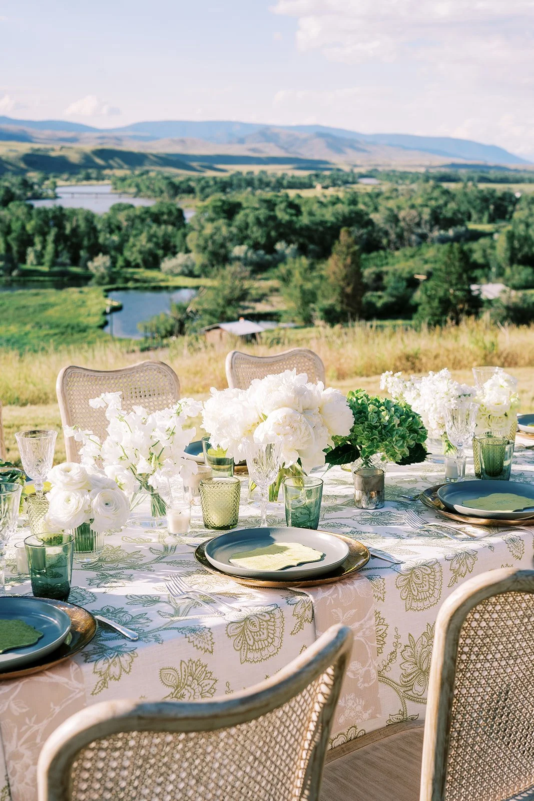 Elegant outdoor dining table set with white floral arrangements, glassware, and plates, overlooking a lush green landscape with hills and a river.