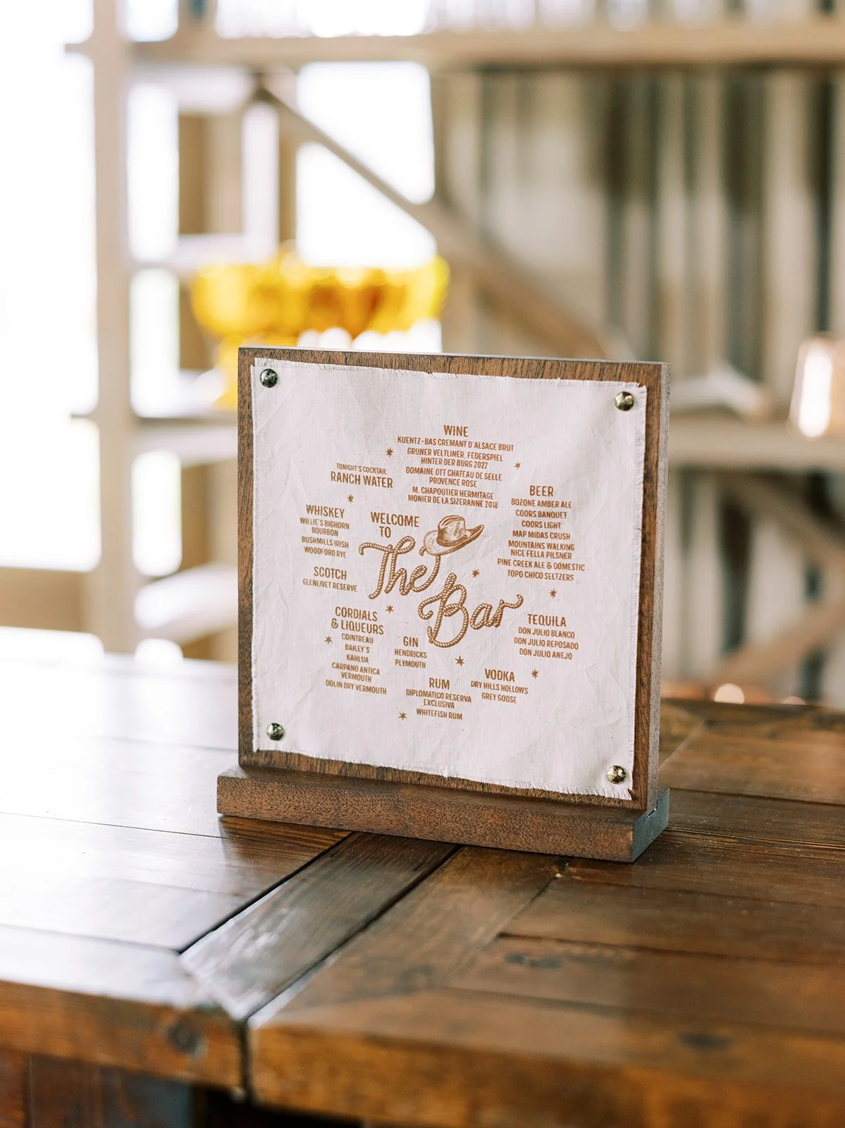 A wooden stand holding a menu titled 'The Bar' with various drink categories and options on a wooden table in a cozy indoor space with blurred background.