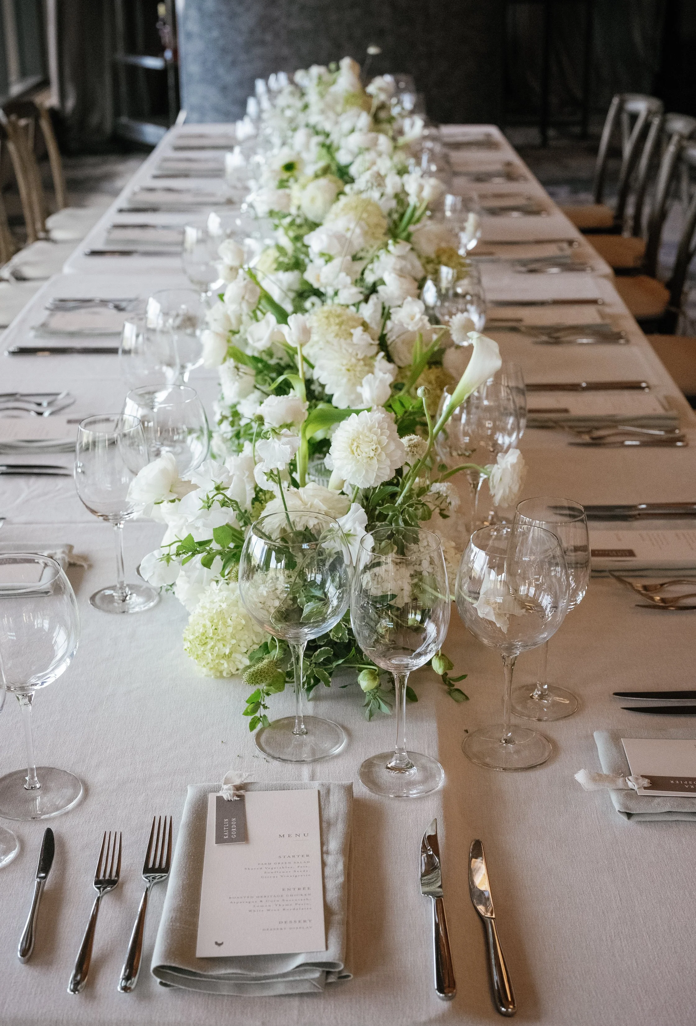Elegant dining table set with white tablecloth, floral centerpiece with white flowers, wine glasses, silverware, and a printed menu card at each place setting.
