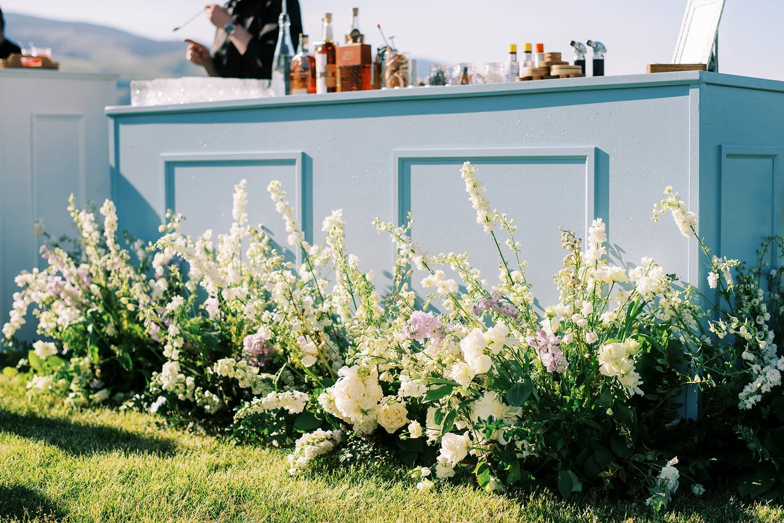 A light blue bar or counter with bottles of liquor and glassware on top, set outdoors, with a flower bed of white and pink flowers in front.
