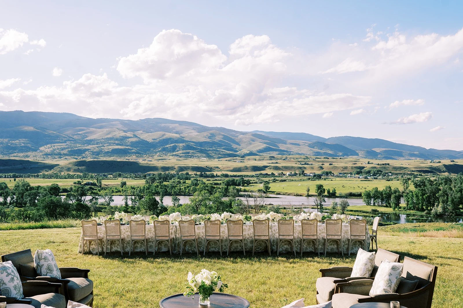 Outdoor event setup with a long banquet table and chairs, overlooking a river and rolling hills under a partly cloudy sky.