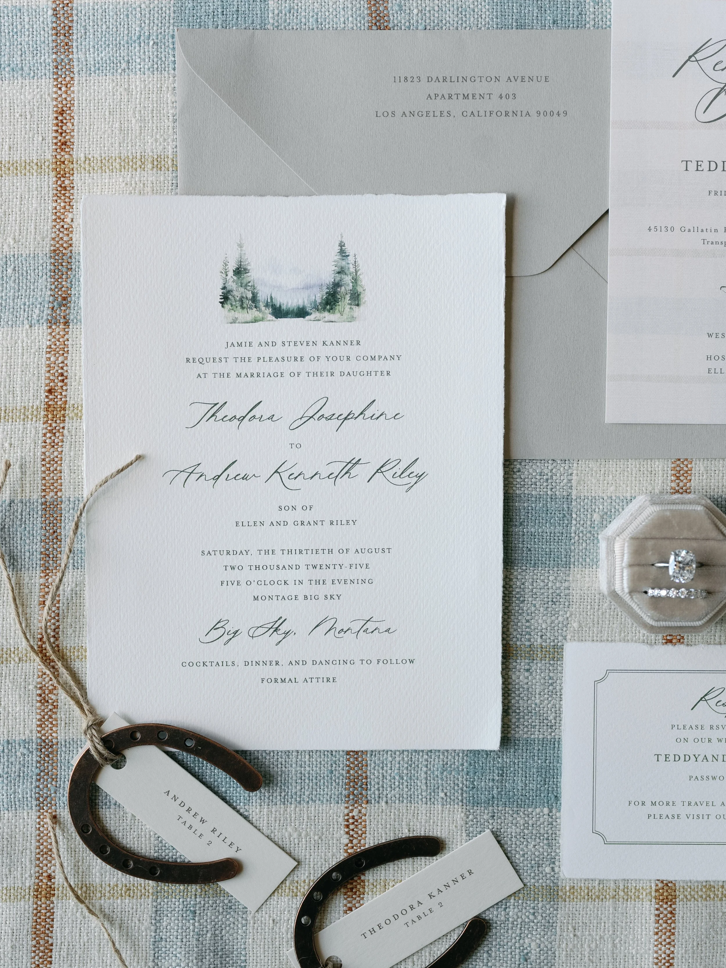 Wedding invitation suite with a white card featuring a watercolor forest scene and elegant script text, along with gray envelopes, horse shoes, and small cards on a plaid fabric background.