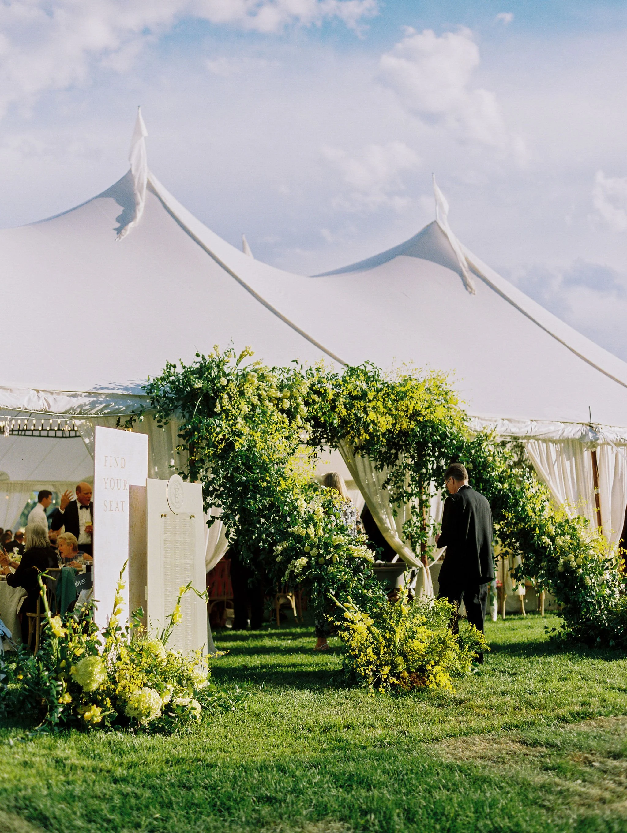 Lush greenery and floral arch framing the entrance to a white tented wedding venue in an outdoor Montana setting, by Wild Blume