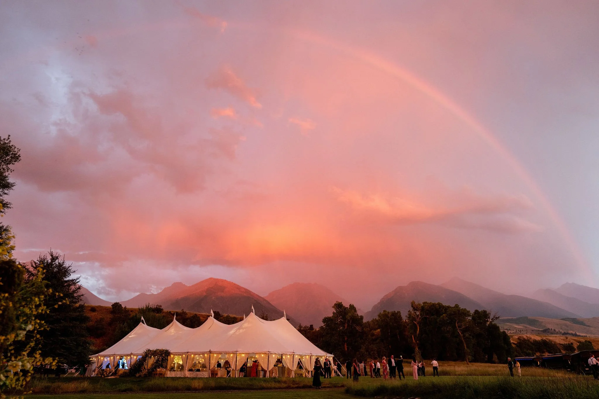 A large white event tent with open sides, set up outdoors at sunset with pink and orange sky, surrounding trees, mountains in the background, and people gathered underneath and nearby.