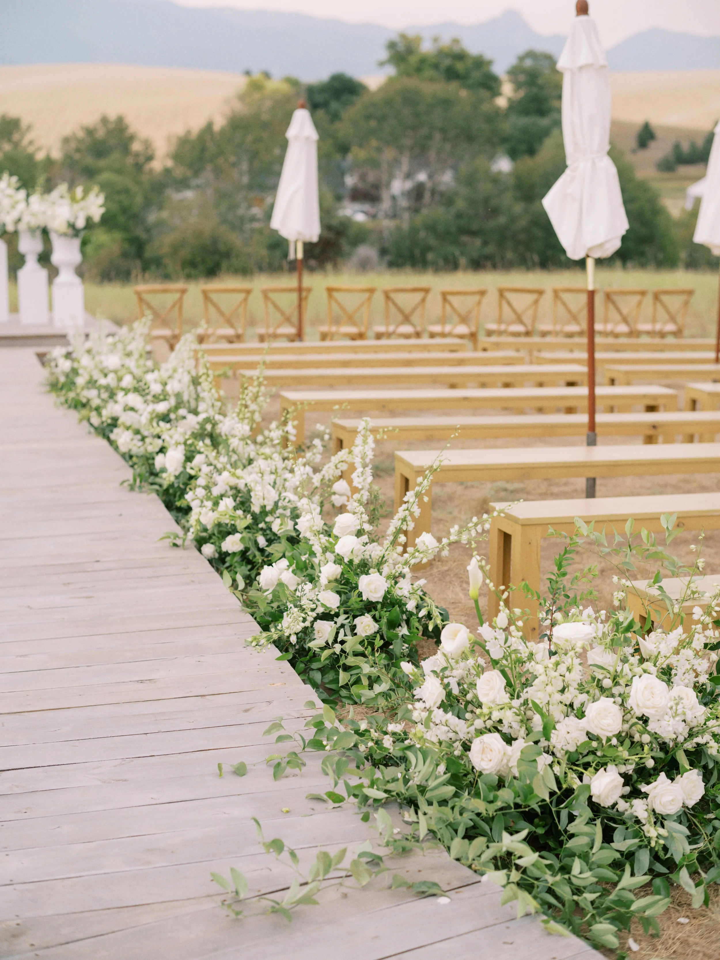 Outdoor wedding ceremony setup with wooden benches, white umbrellas, and floral arrangements in a rural landscape.